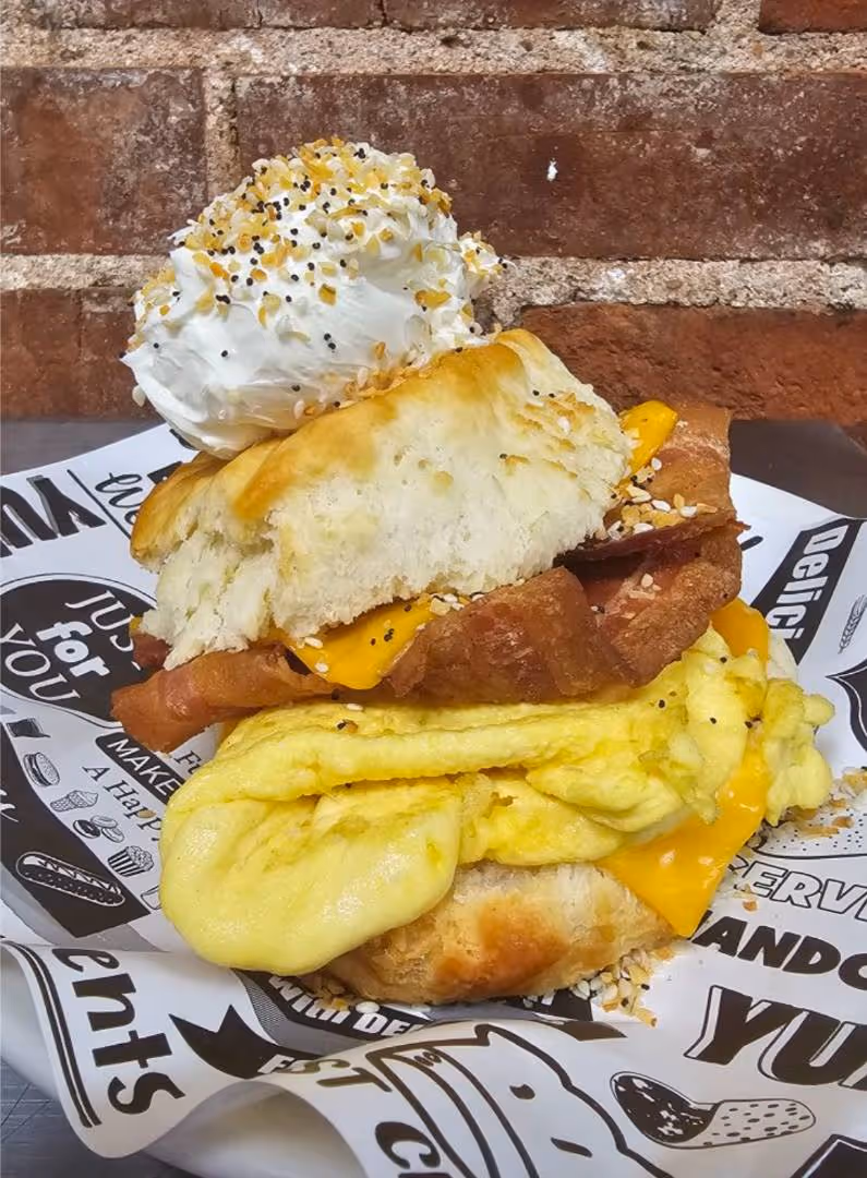 Breakfast sandwich with scrambled eggs, cheddar cheese, and bacon between biscuits, topped with cream cheese and seasoning, on graphic paper. Brick wall backdrop.