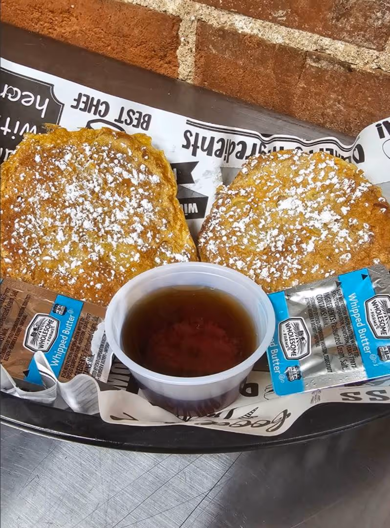 Two french toast biscuits dusted with powdered sugar, served in a basket with syrup and packets of whipped butter on a paper-lined tray.
