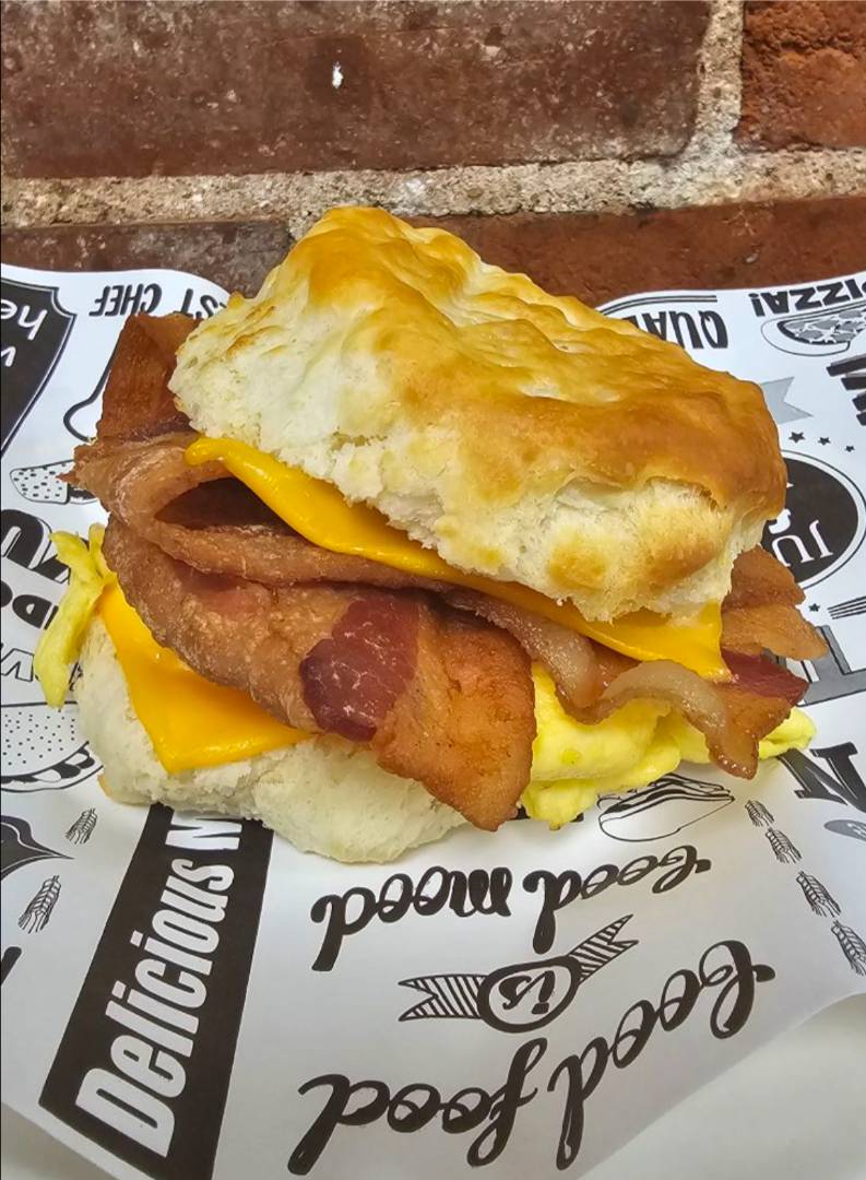 A savory breakfast biscuit sandwich with crispy bacon, melted cheddar cheese, and fluffy scrambled eggs, set on branded paper against a brick wall.