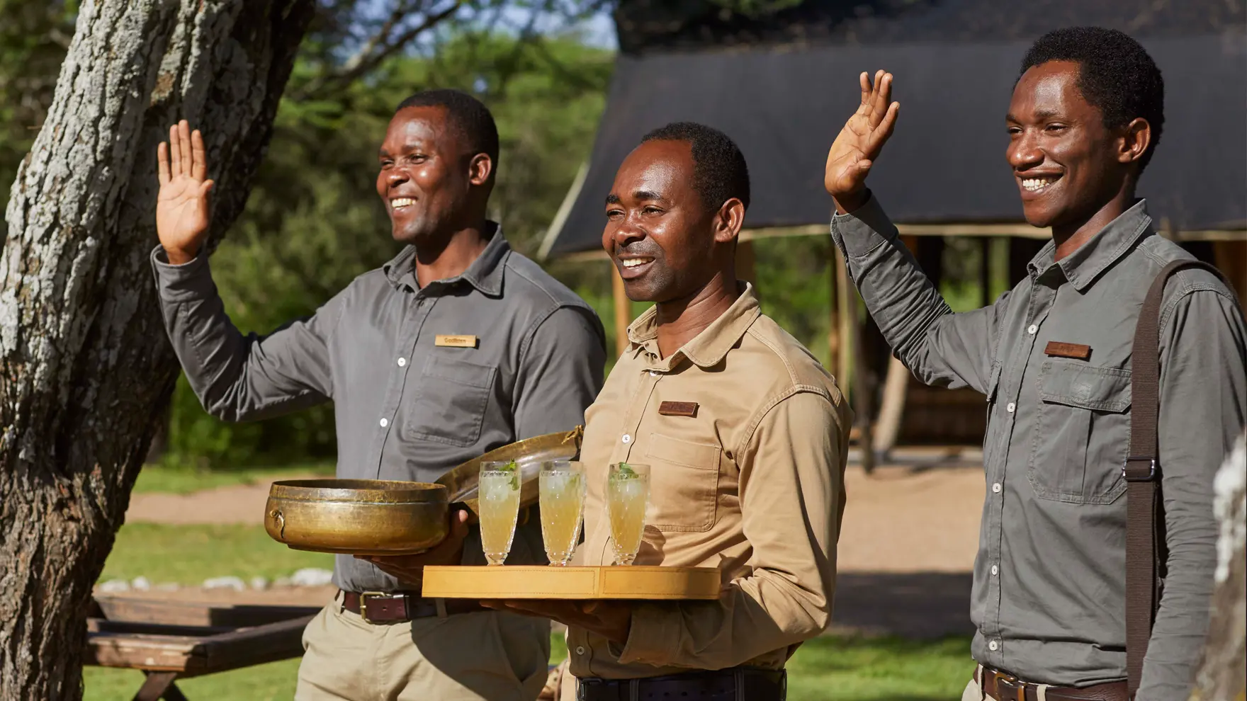 Three smiling staff members greeting guests, holding beverages and a welcome gong at a safari camp.