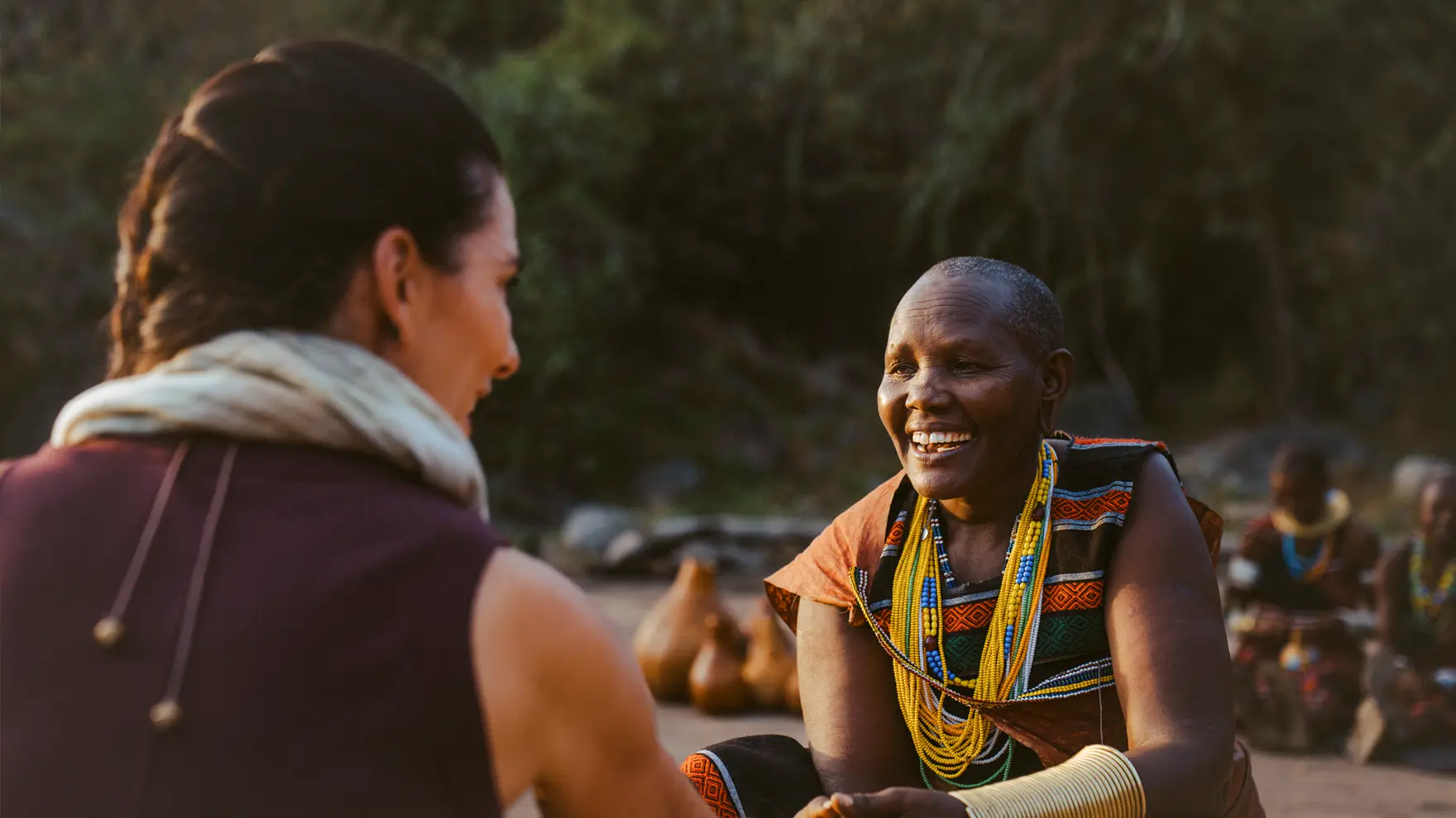 Smiling local woman wearing colourful beaded jewellery engaging warmly with a visitor in an outdoor cultural setting.