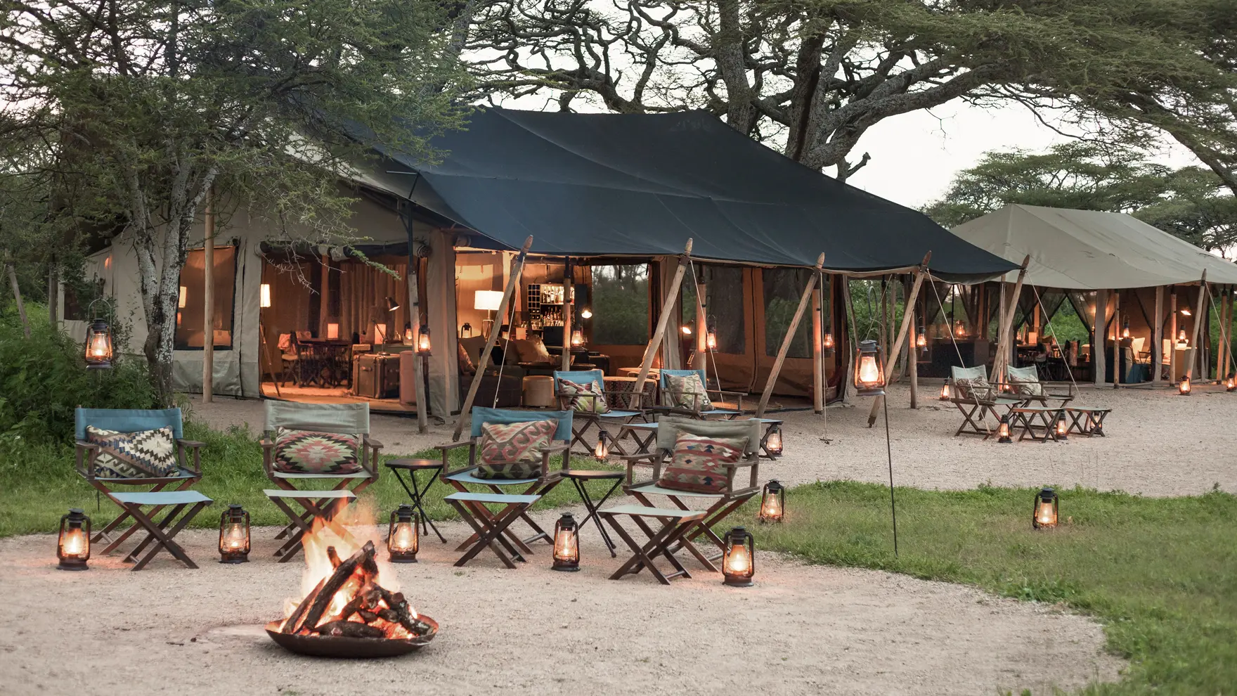 Open-air safari camp lounge at dusk with canvas tents, lanterns, wooden chairs, and a glowing fire pit.