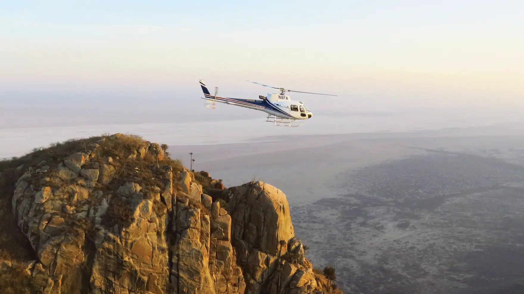 Helicopter flying above a rugged rocky cliff at sunrise with expansive plains stretching into the distance.