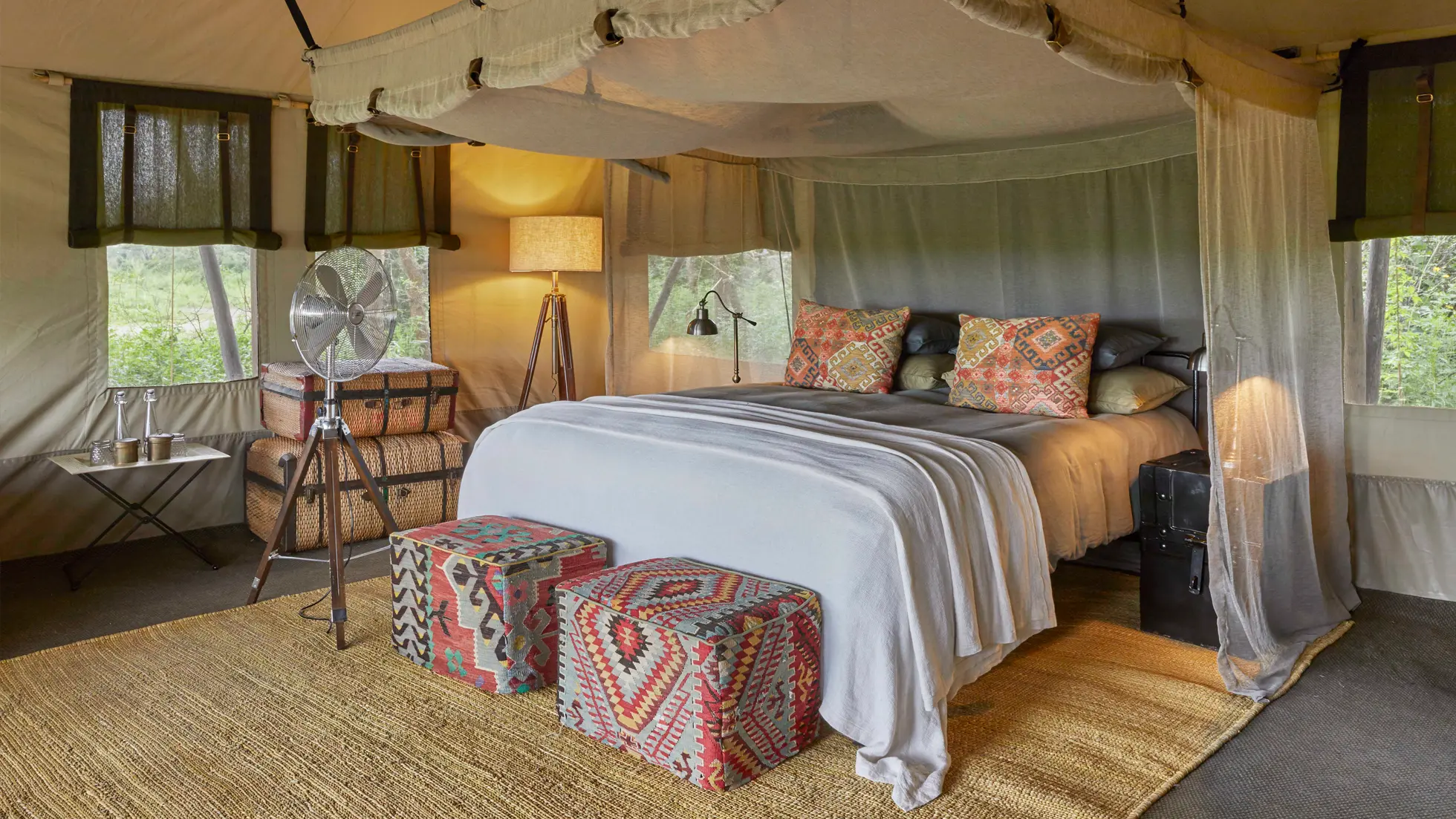 Interior of a stylish tented camp bedroom with a made bed, patterned cushions, two colorful ottomans, a tripod fan, and a wicker trunk.