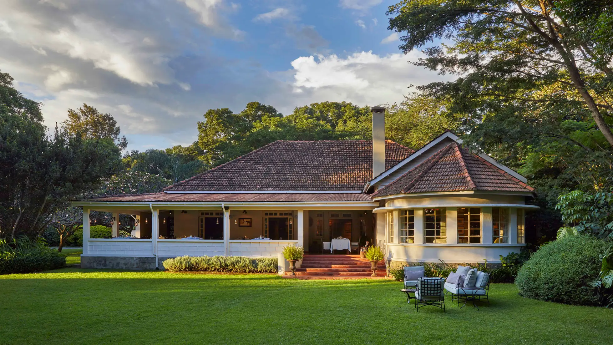 Elegant lodge with a tiled roof surrounded by lush trees and a green lawn with outdoor seating.