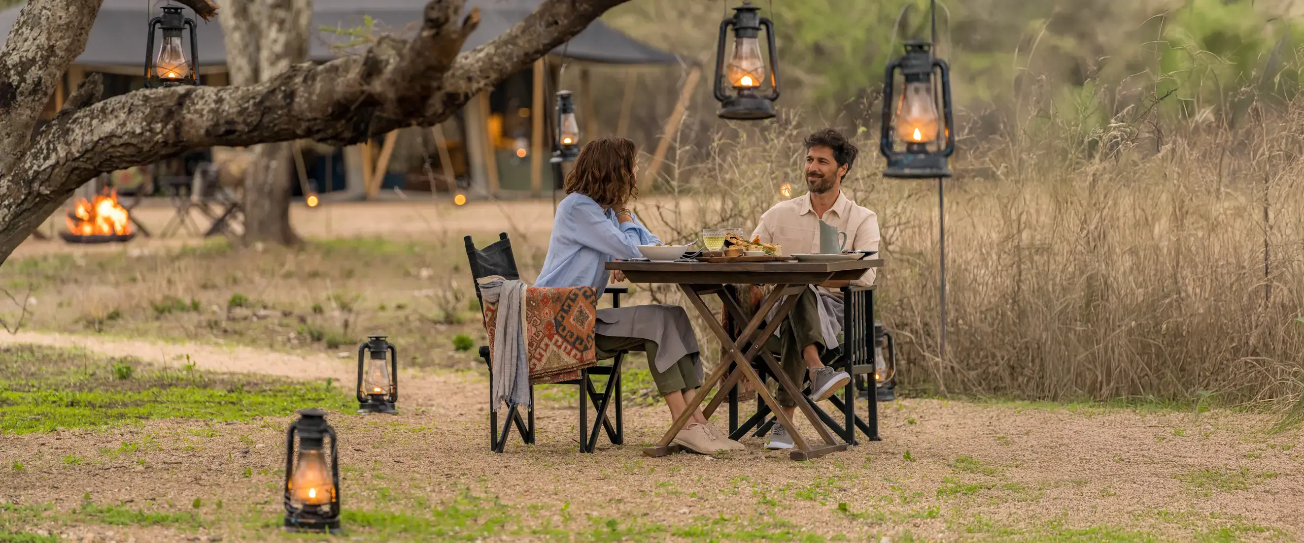 Man and woman sitting at a wooden table outdoors having a meal surrounded by lit lanterns hanging and on the ground.