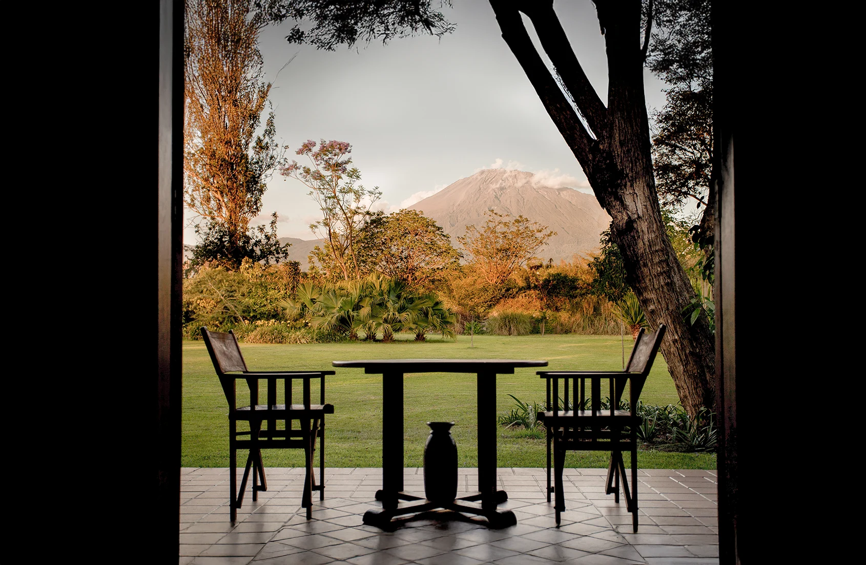 Two wooden chairs and a table on a tiled patio overlooking a grassy lawn with trees and a mountain in the background during sunset.