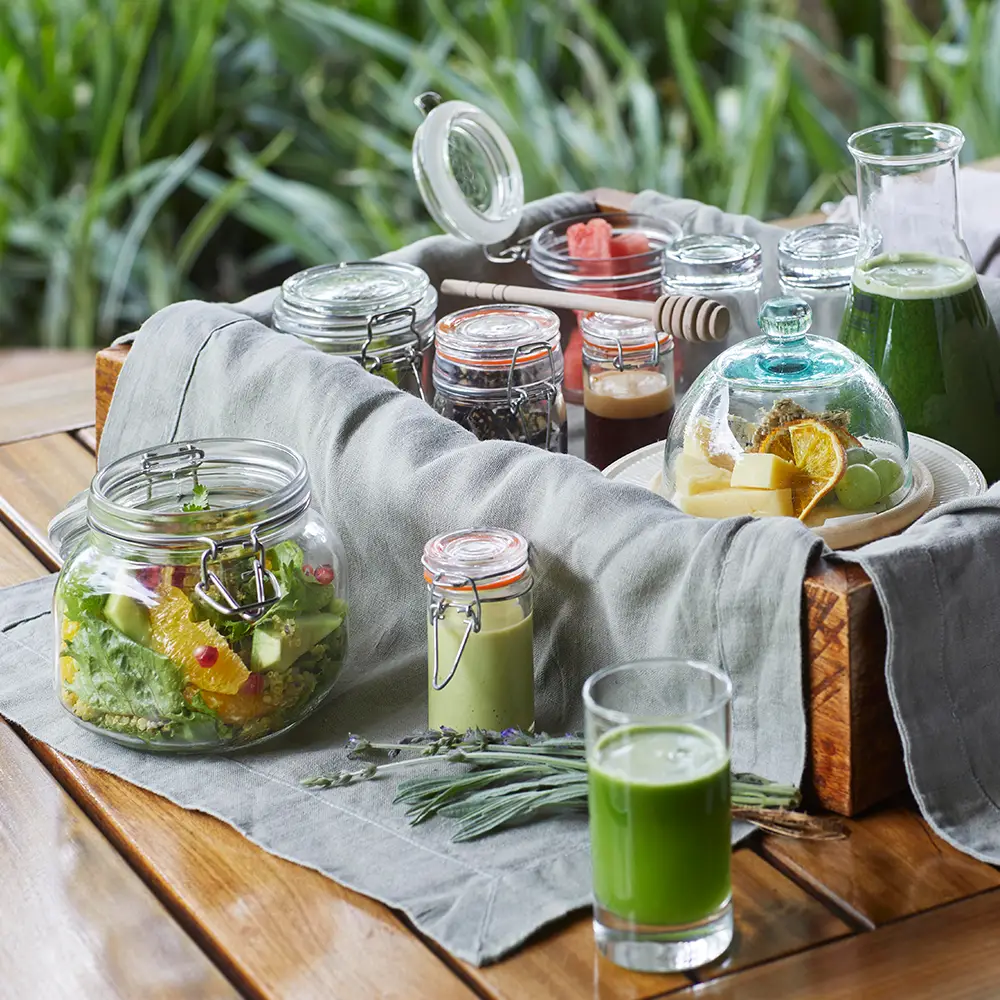 Wooden tray with jars of salad, green juice, honey dipper, and snacks on a gray cloth on a wooden table outdoors.