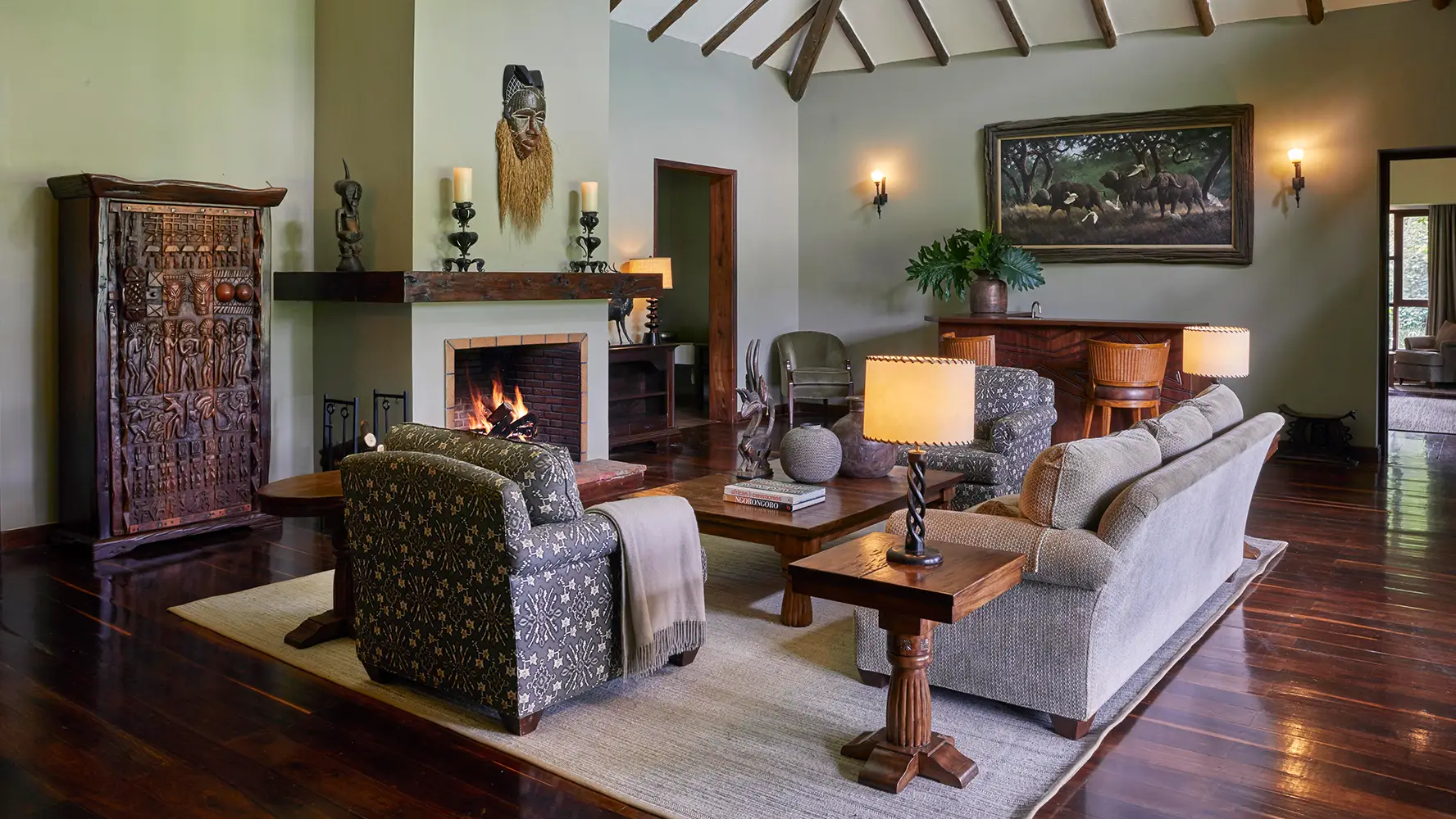 Cozy living room with wooden floor, a lit fireplace, patterned armchairs, a beige sofa, wooden tables with lamps, tribal mask above the mantle, and a painting of elephants on the wall.