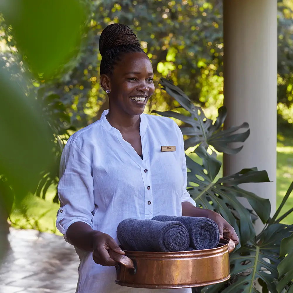 Smiling woman in a white shirt holding a copper tray with two rolled gray towels outdoors.