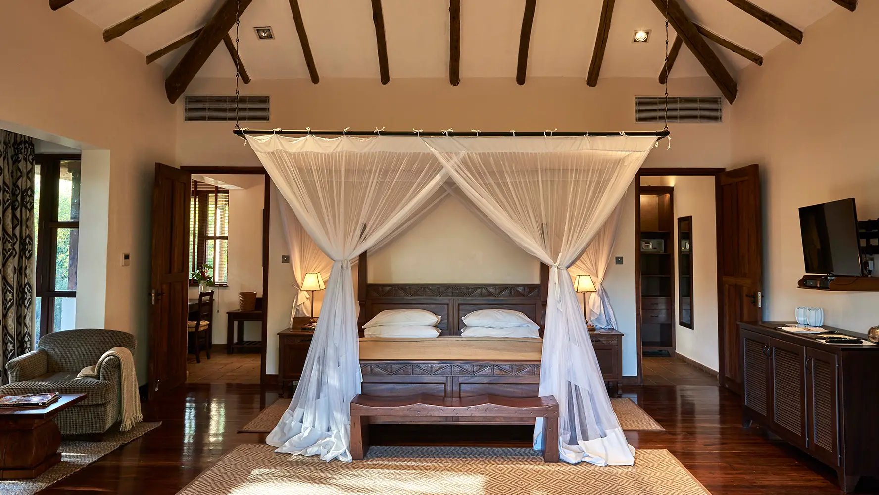 Spacious bedroom with a wooden four-poster bed draped with white sheer curtains, flanked by bedside tables with lamps, and wooden furniture on polished hardwood floors.