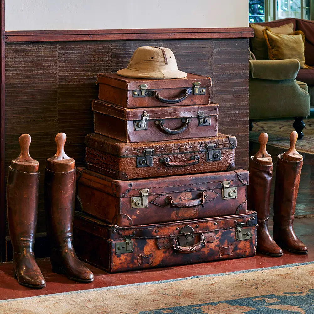Stack of five vintage brown leather suitcases topped with a beige safari hat, flanked by two pairs of tall brown leather riding boots with wooden boot trees inside.