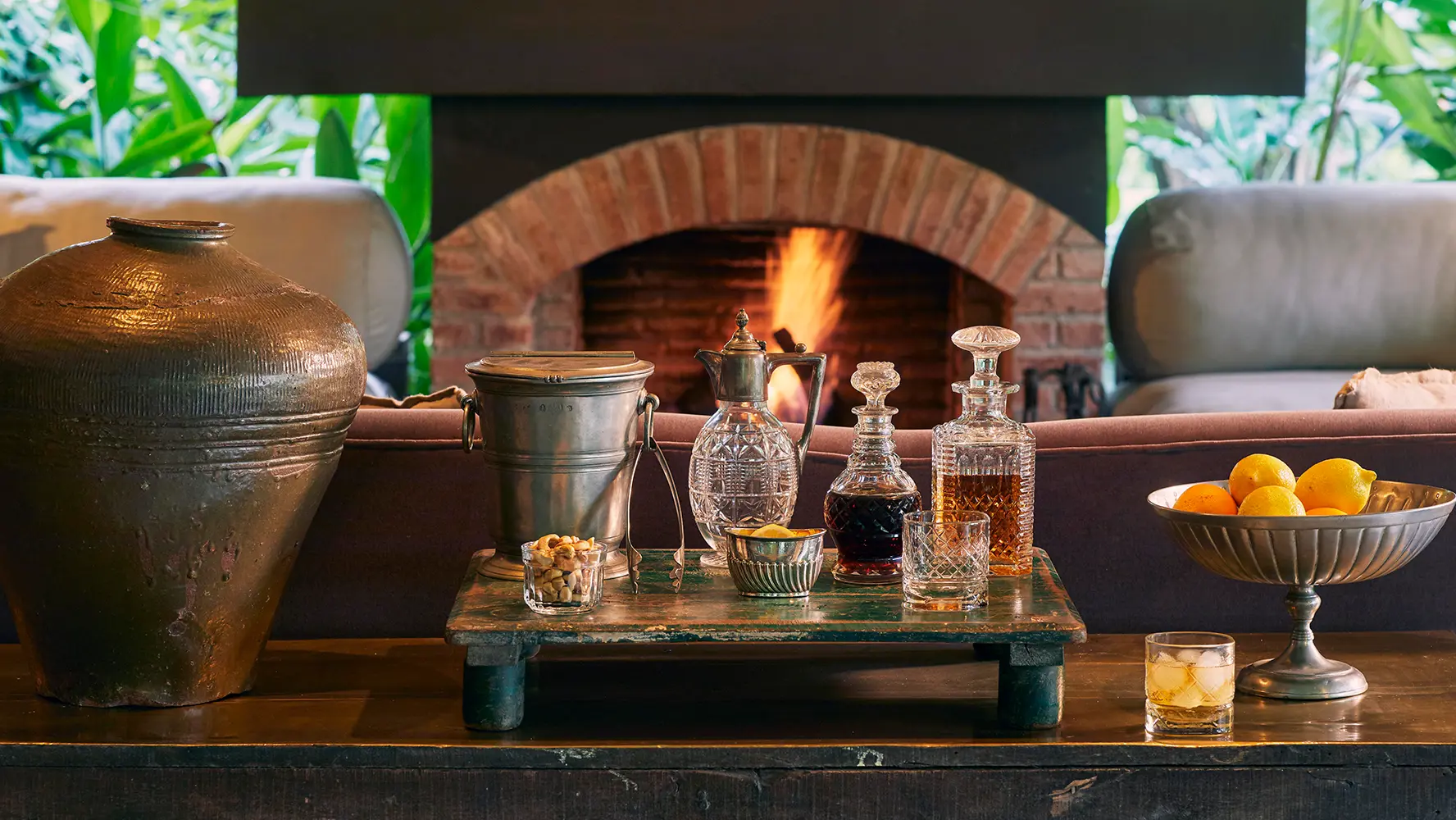 Rustic indoor setting with a lit brick fireplace, a wooden table holding assorted glass decanters, a metal ice bucket, a bowl of mixed nuts, a bowl of lemons and oranges, and a glass of whiskey.