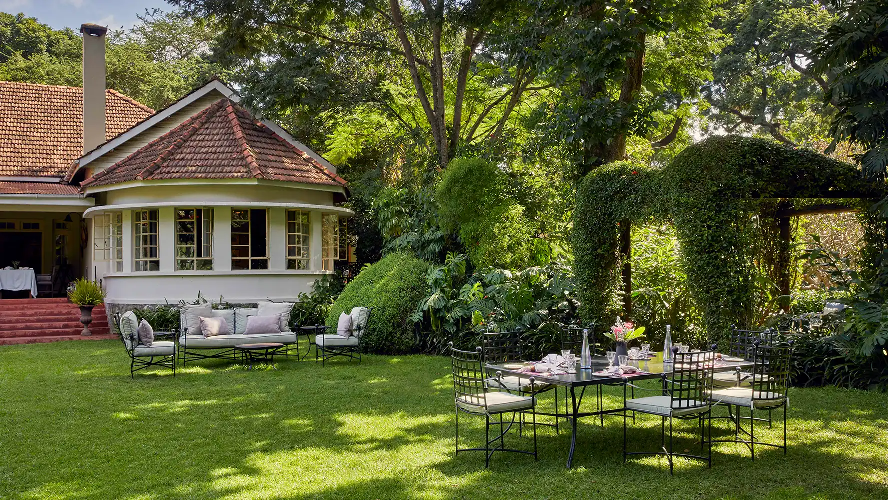 Outdoor garden seating area with a cushioned sofa set on green lawn beside a round table with chairs under lush greenery and a vine-covered arch.