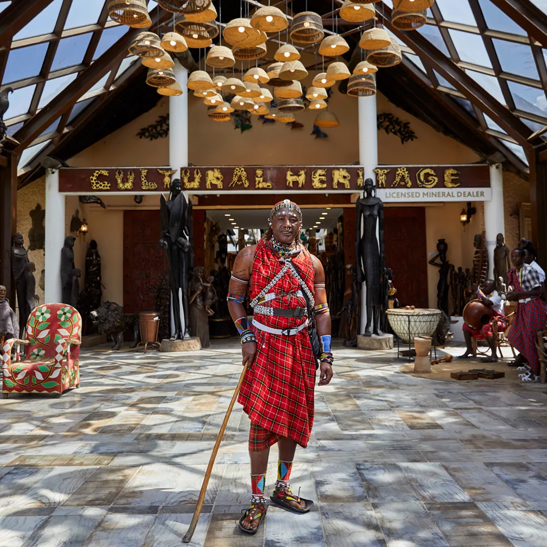 Man in traditional Maasai attire holding a wooden staff, standing inside a cultural heritage center with hanging basket lamps and wooden sculptures.