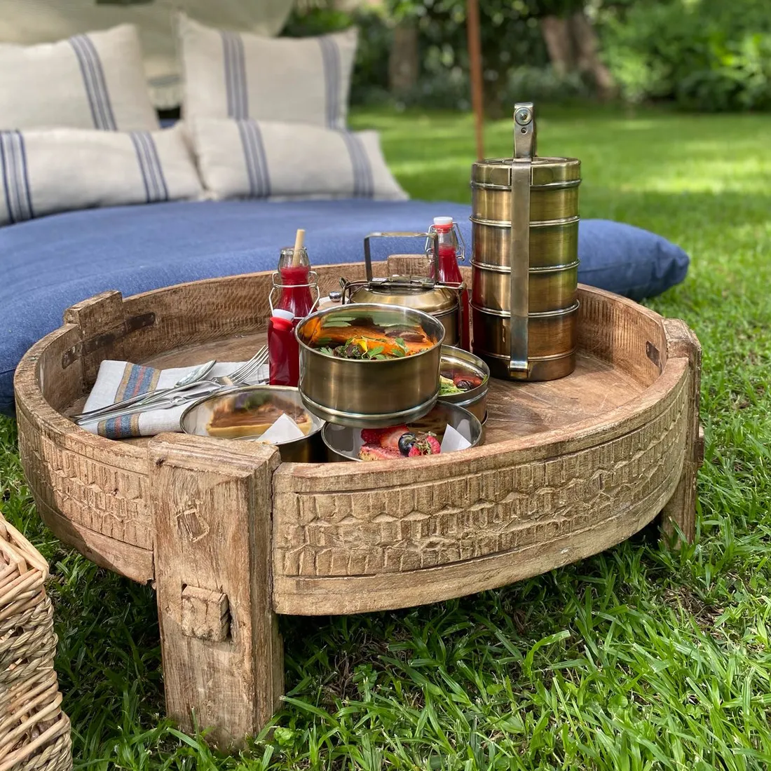 Wooden tray on grass holding stacked food containers, with a cushioned outdoor seating in the background.