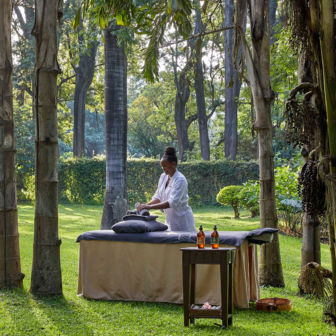 Woman in white attire arranging towels on a massage table set outdoors in a lush green garden with trees.