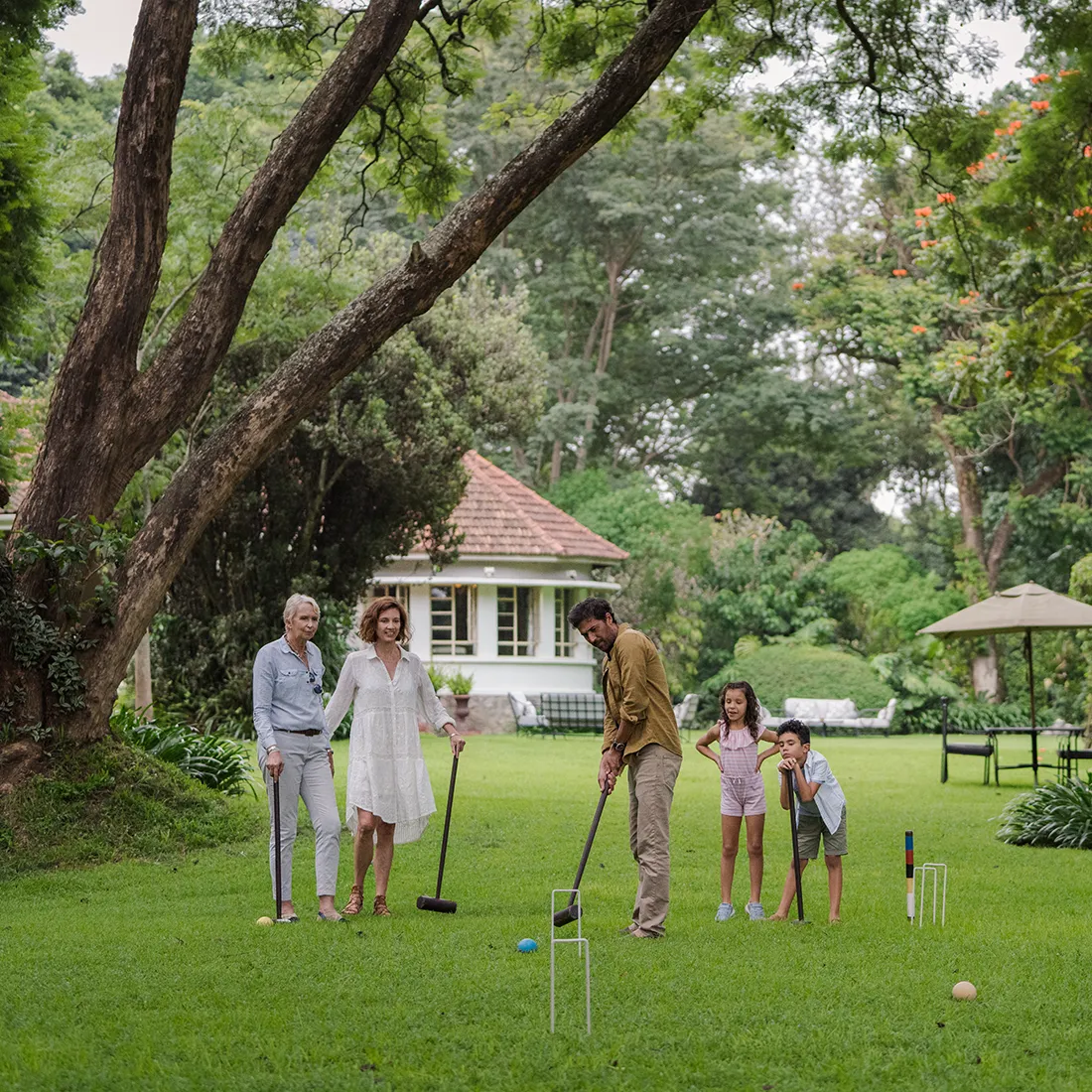 A family playing croquet on a green lawn with trees and a house in the background.