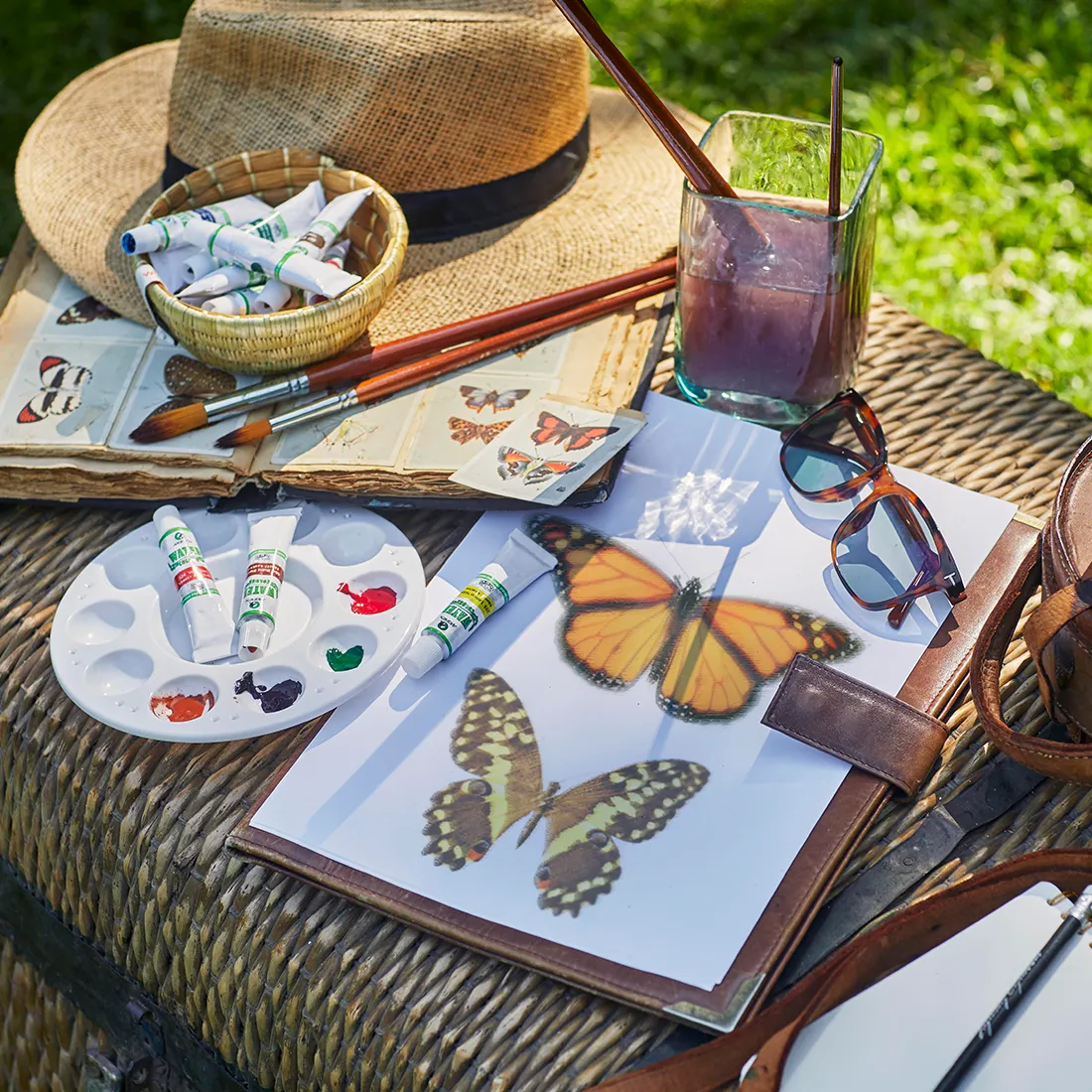 Table with butterfly illustrations, watercolors, paintbrushes, sunglasses, and a straw hat on a sunny day.