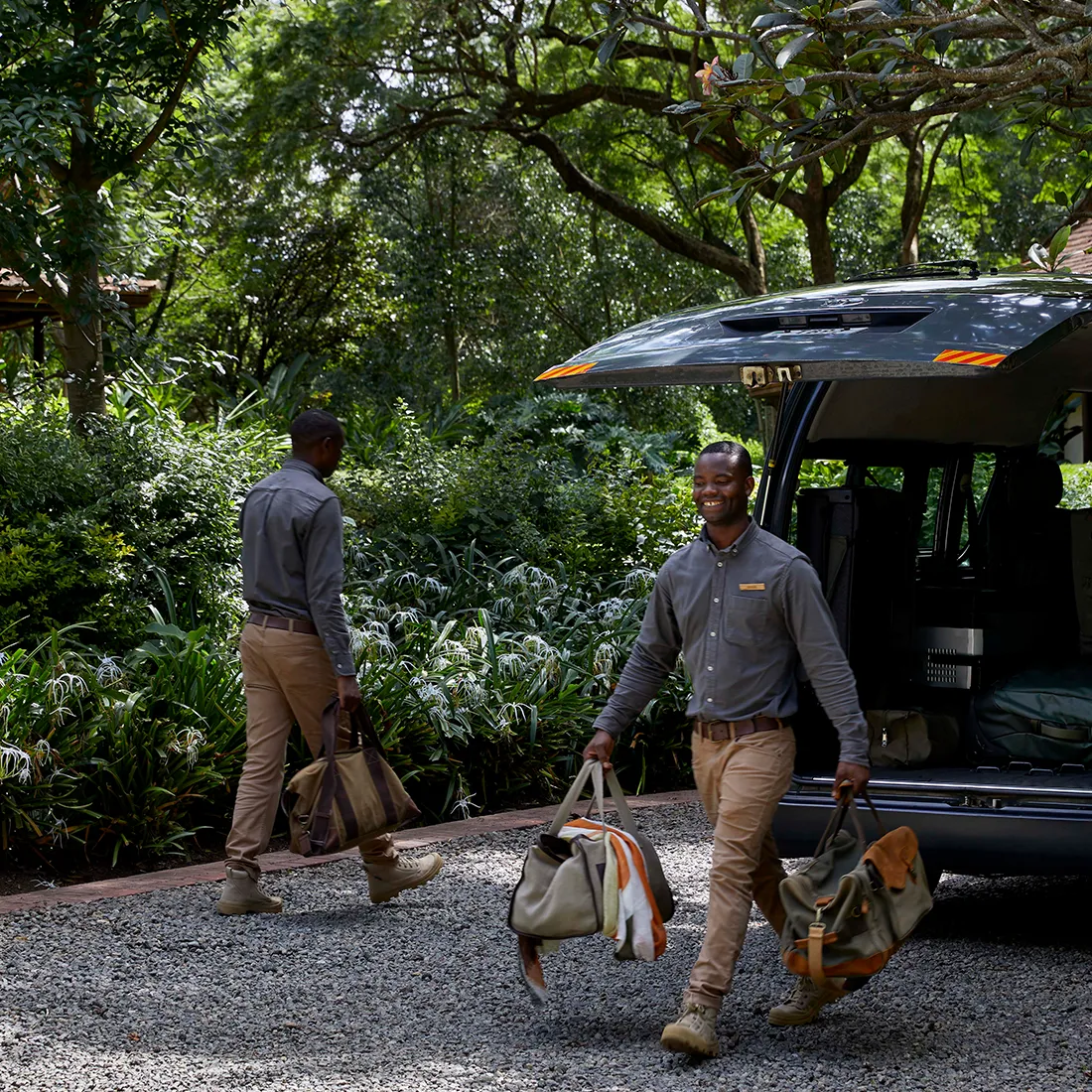 Two men carrying duffel bags walking away from an open vehicle trunk in a lush green outdoor setting.