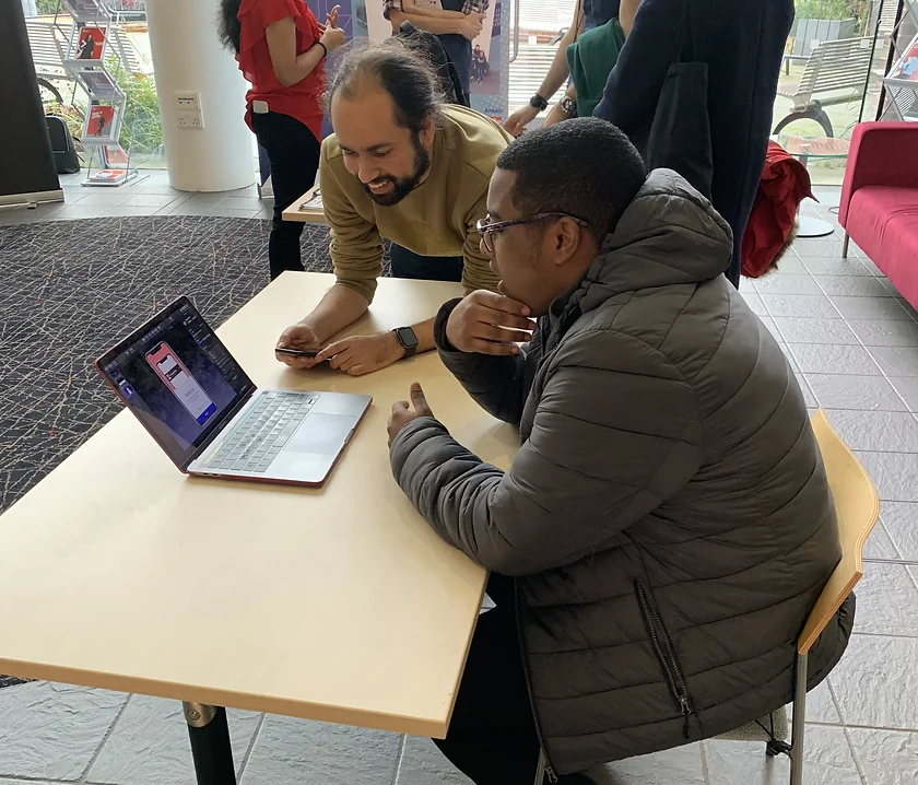 Two men engaged in a discussion while looking at a laptop on a light wooden table in a modern indoor setting.