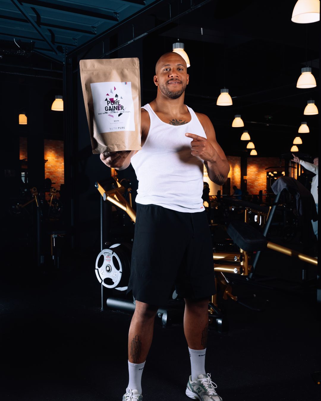 Homme musclé en débardeur blanc tenant un sac de poudre protéinée Pure Gainer en salle de sport.