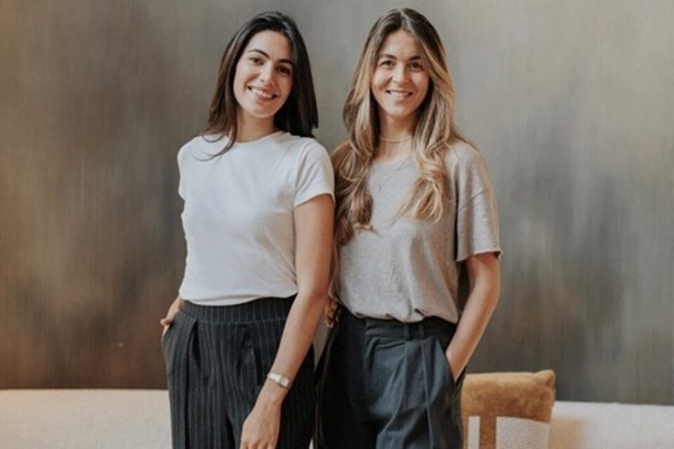 Deux femmes souriantes debout côte à côte, mains dans les poches, devant un mur gris.