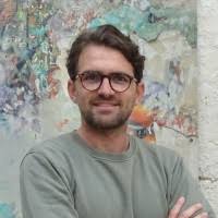 Un homme portant des lunettes, un t-shirt vert, bras croisés, souriant devant un mur peint.