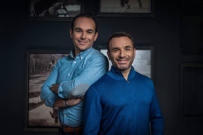 Deux hommes souriants, l'un en chemise bleue claire, l'autre en pull bleu foncé, posant devant un mur sombre.