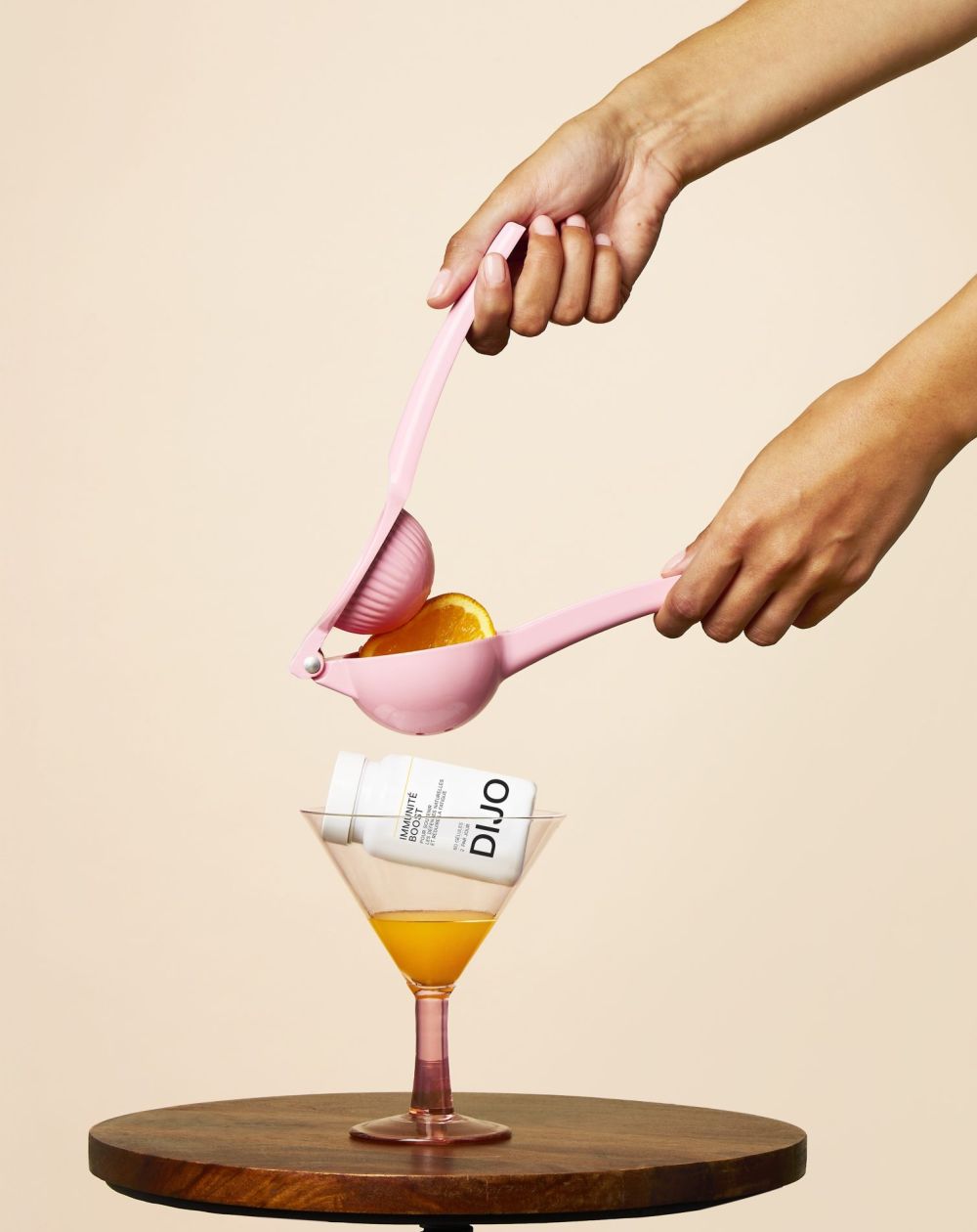 Deux mains pressent une demi-orange avec un presse-agrumes rose au-dessus d'un verre à cocktail avec une bouteille DIJO.