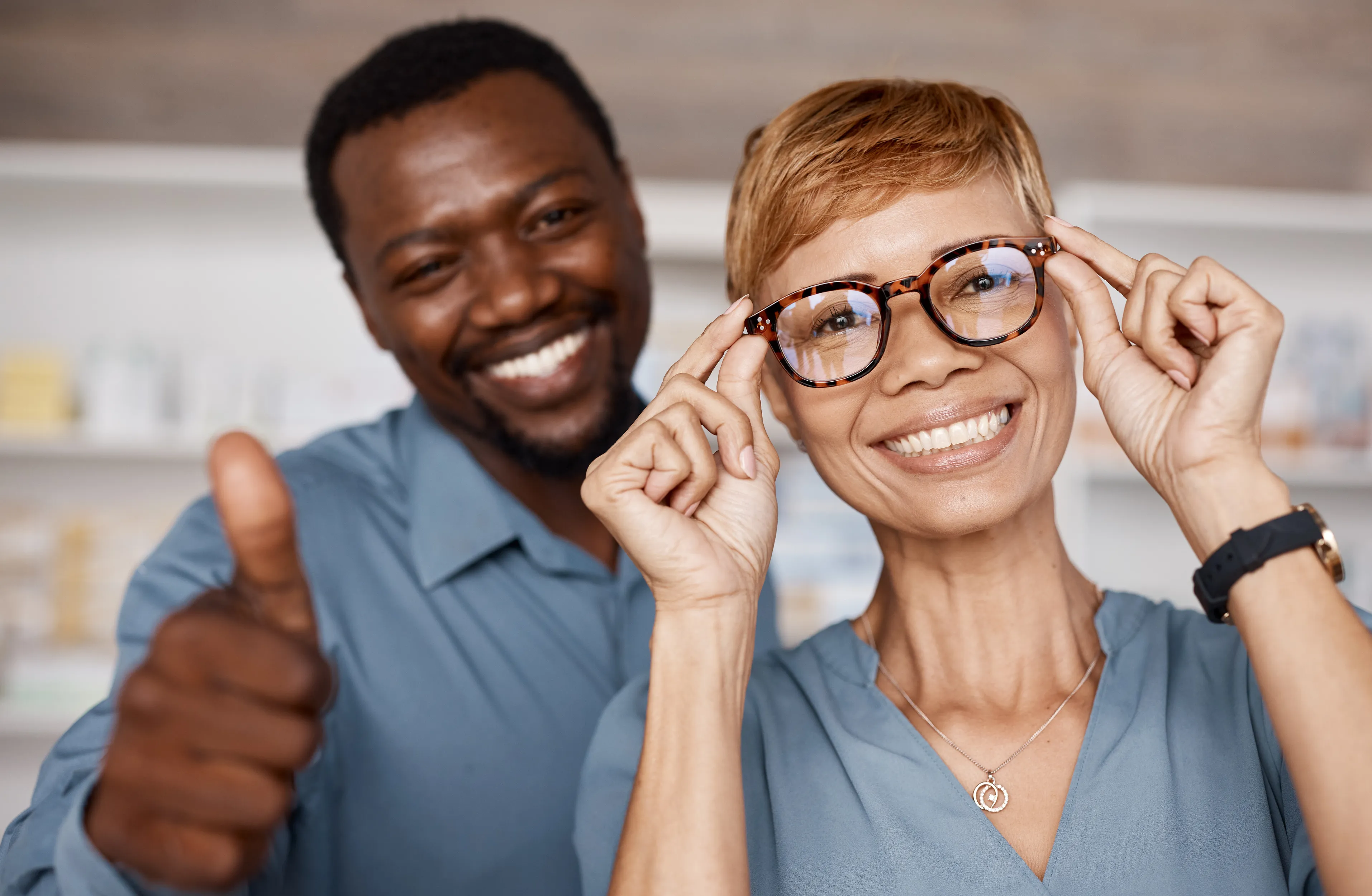 Two coworkers in blue shirts smiling, one giving thumbs up, adjusting glasses