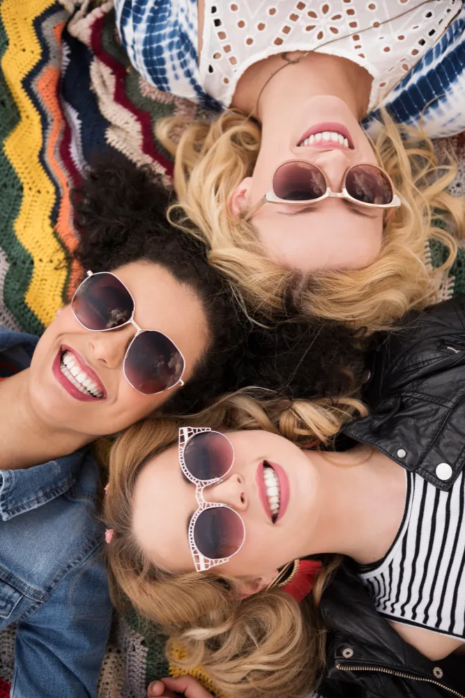 Three friends lying down wearing sunglasses and smiling on colorful blanket