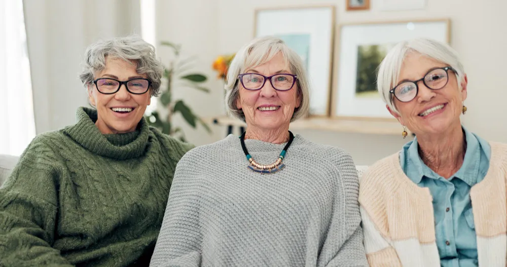 Three senior women smiling together, wearing cozy sweaters and glasses