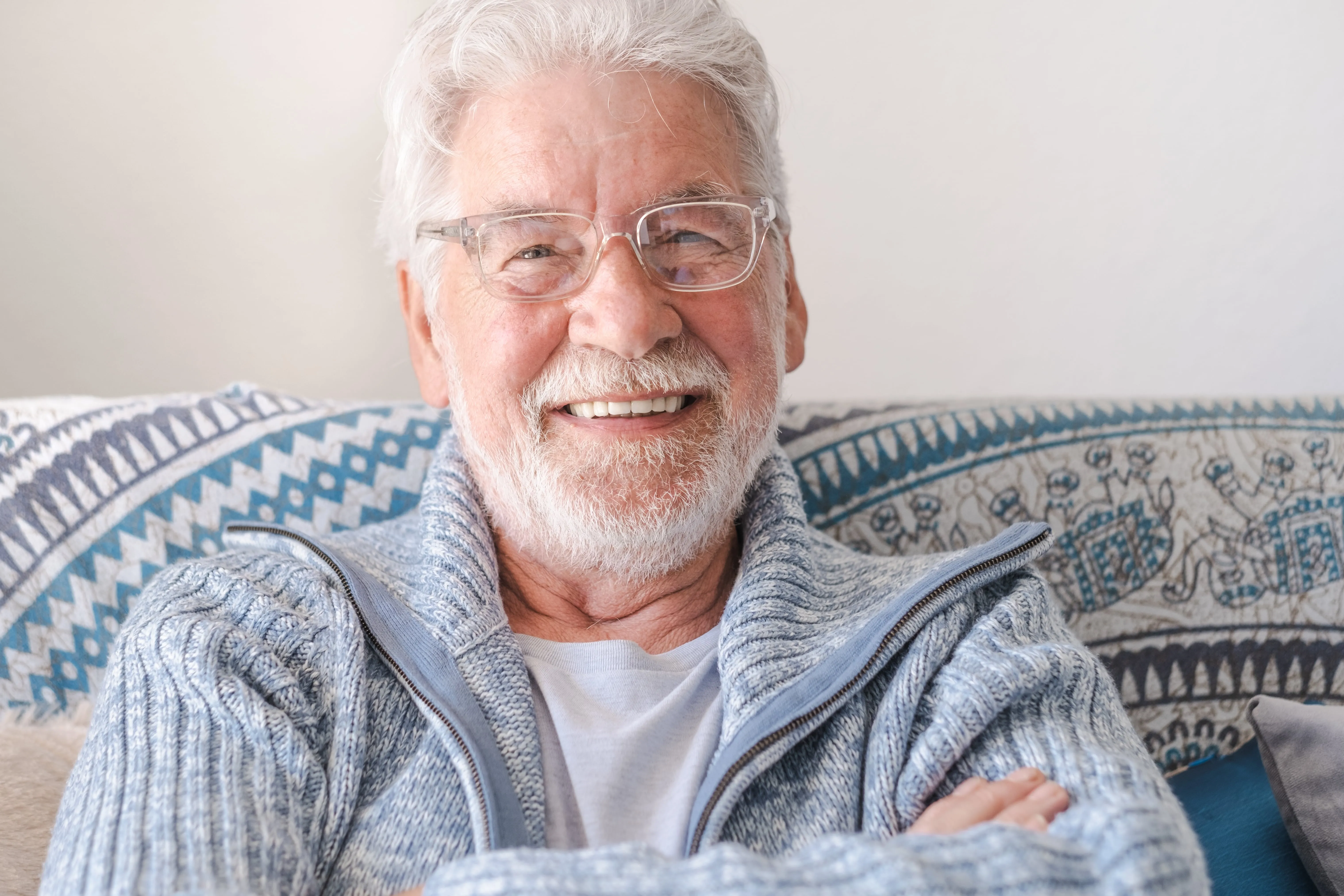 Smiling elderly person in glasses and gray sweater sitting on patterned couch