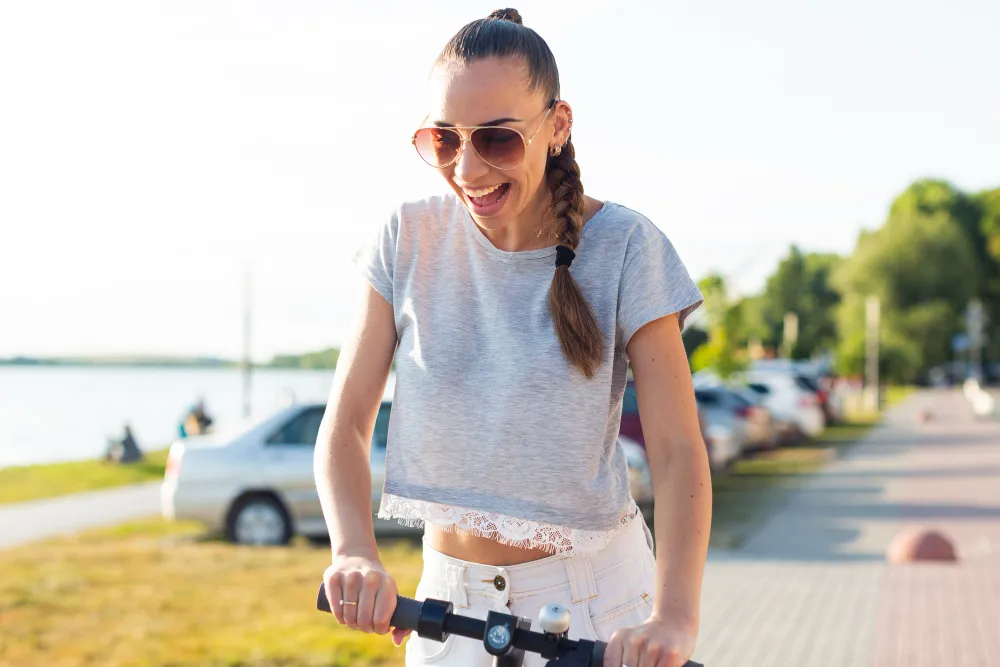 Smiling woman riding electric scooter on sunny street with parked cars
