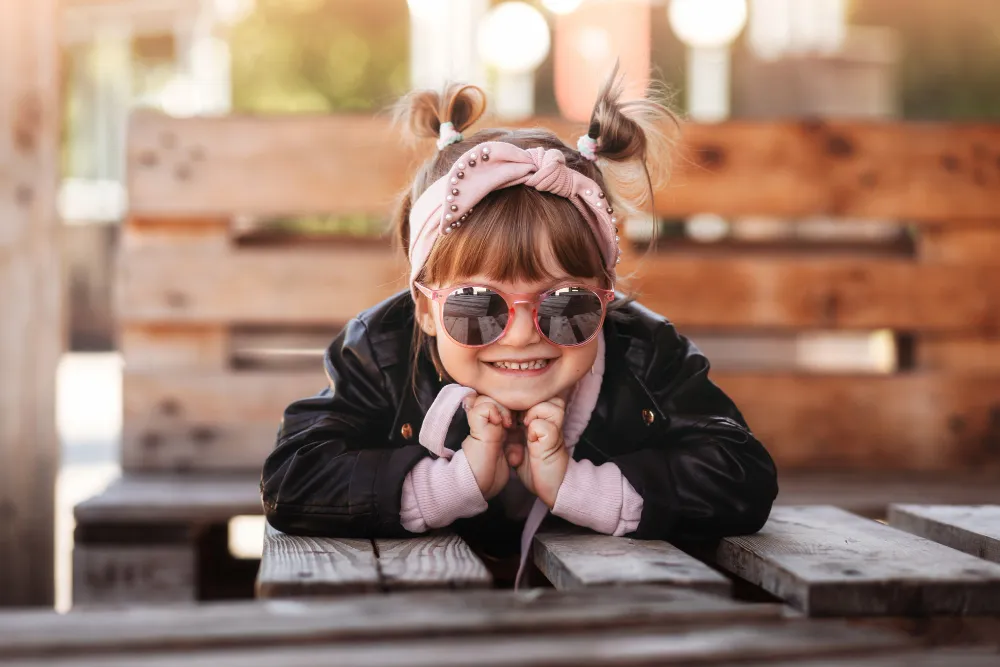 Smiling child with sunglasses and hair bows leaning on wooden bench