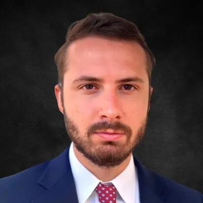 Man with short brown hair and beard wearing a navy suit, white shirt, and red polka dot tie.
