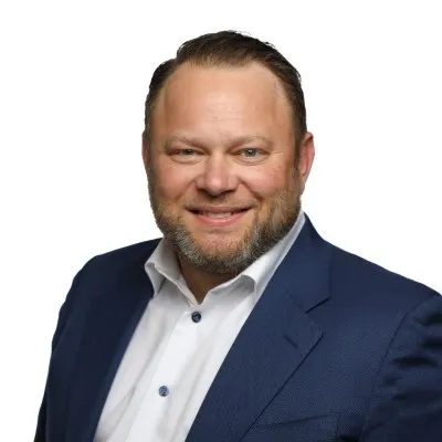 Smiling man with short hair and beard wearing a white shirt and blue suit jacket against white background.