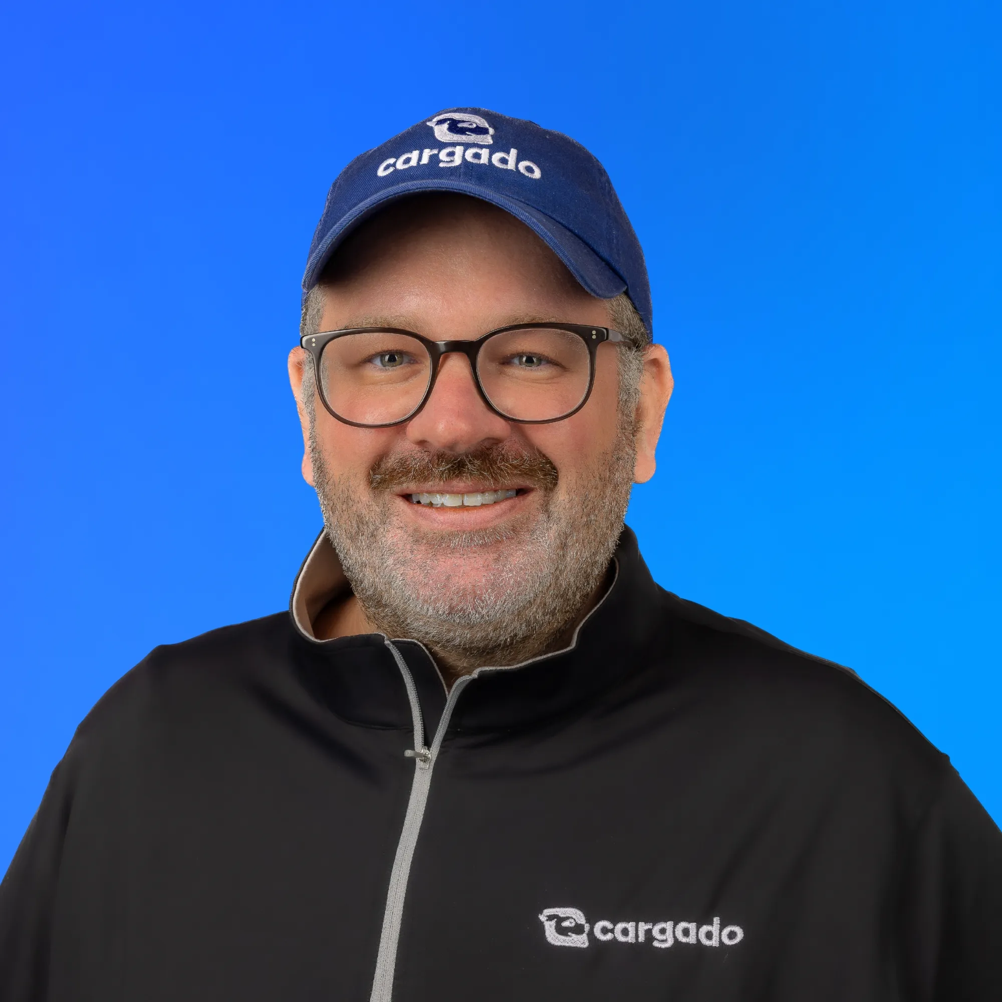 Man with glasses and beard wearing a blue Cargado cap and black jacket smiling on blue background.