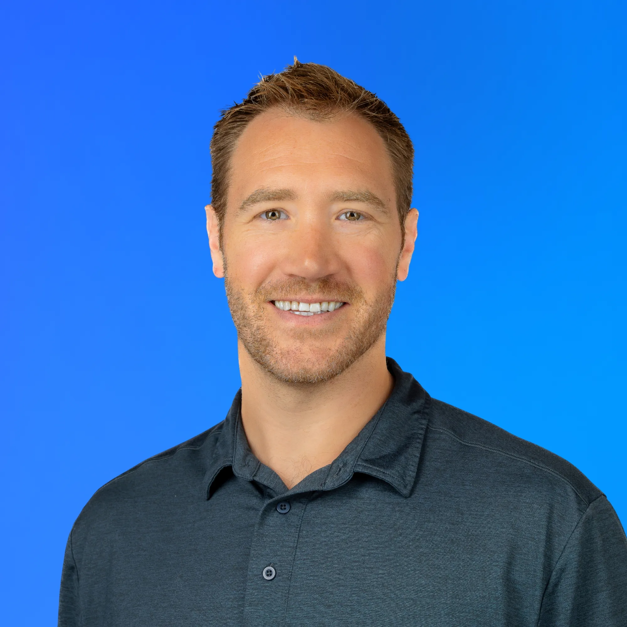 Smiling man with short brown hair and a beard wearing a dark grey collared shirt on blue background.