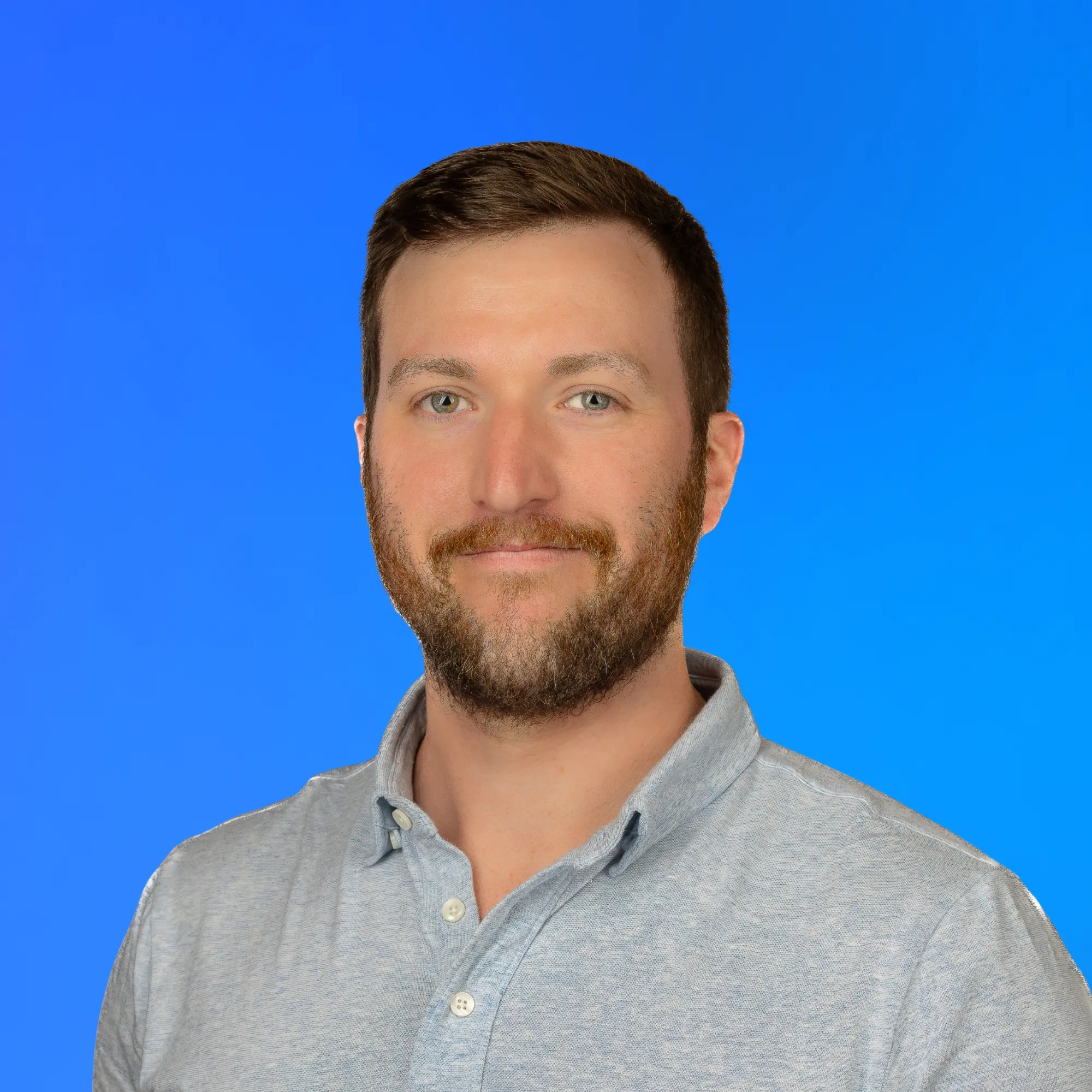 Smiling man with short brown hair and beard wearing a light gray button-up shirt on blue background.