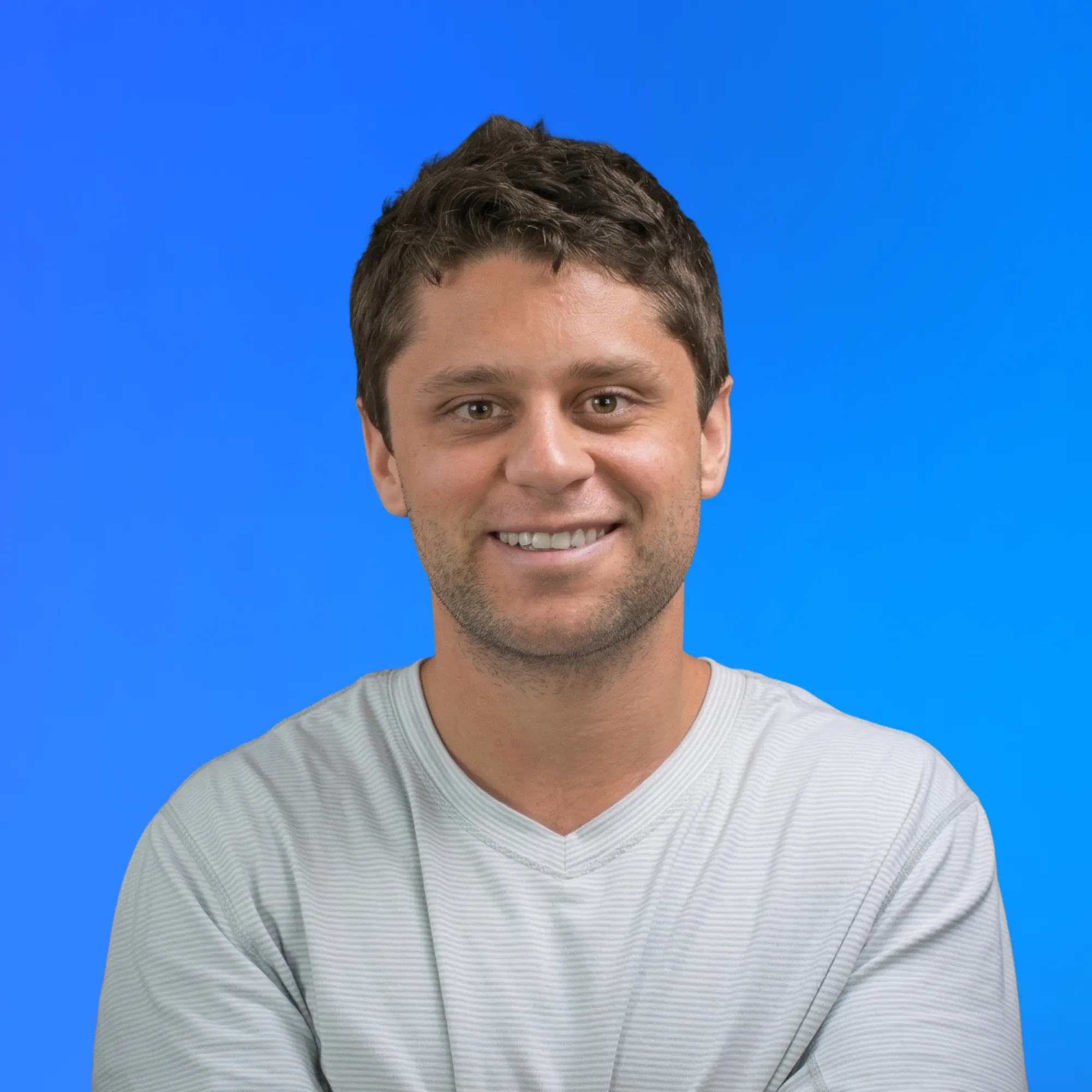Smiling young man with short brown hair wearing a light gray striped shirt against blue background.