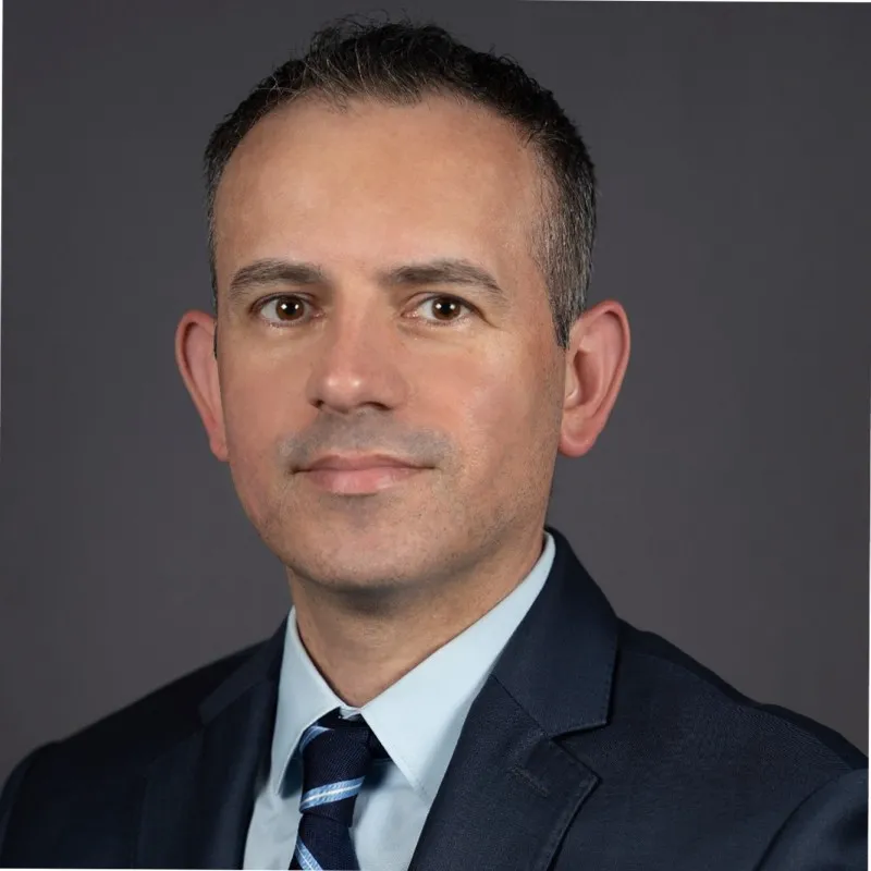 Headshot of a man in a dark suit, light blue shirt, and striped tie against a gray background.