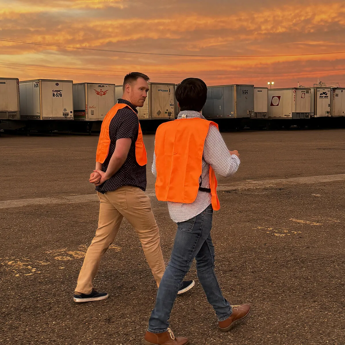Two men wearing orange safety vests walking in a parking lot with trailers at sunset.