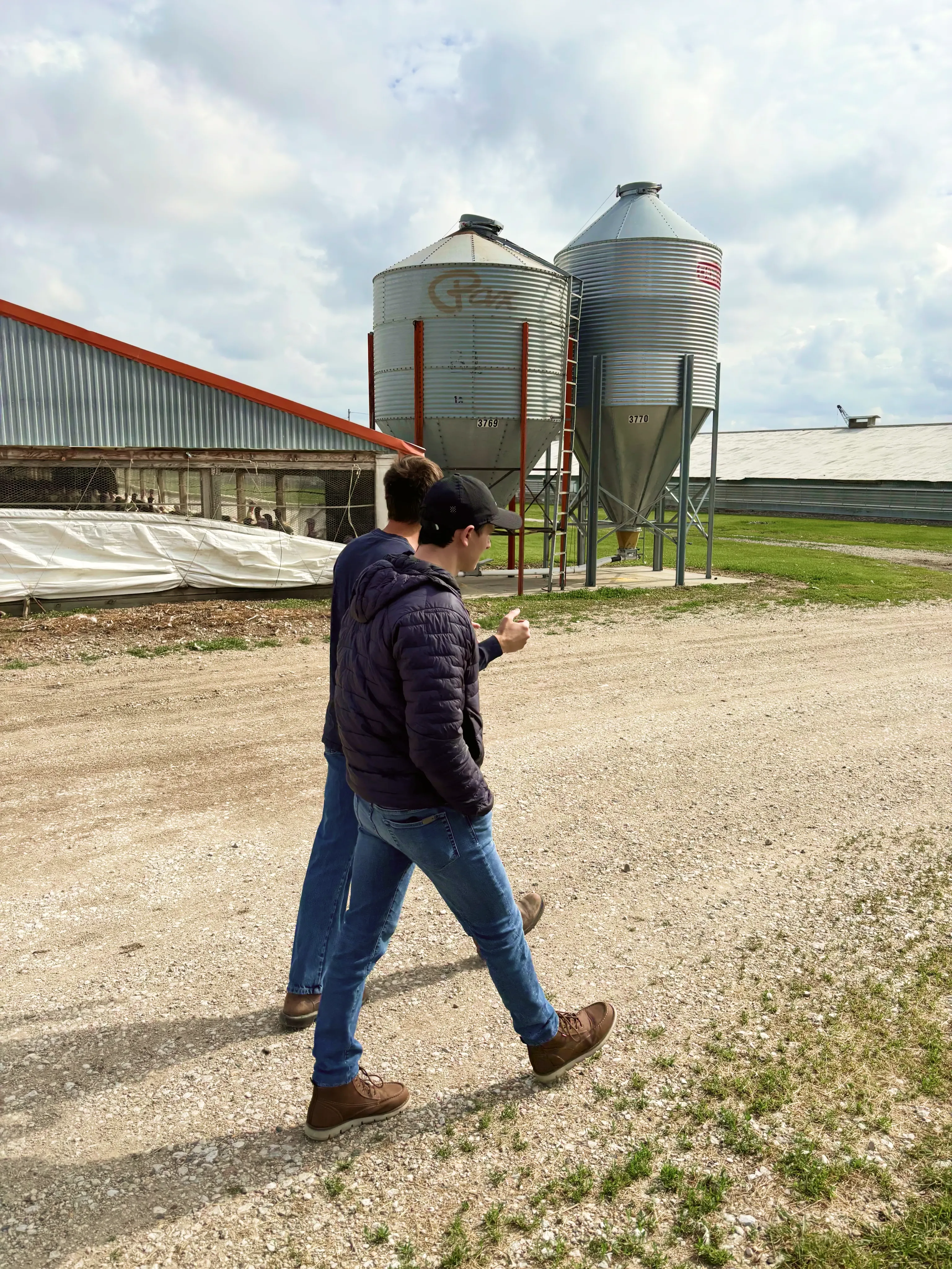 Man in casual clothing walking on dirt road near farm with metal silos and buildings nearby.