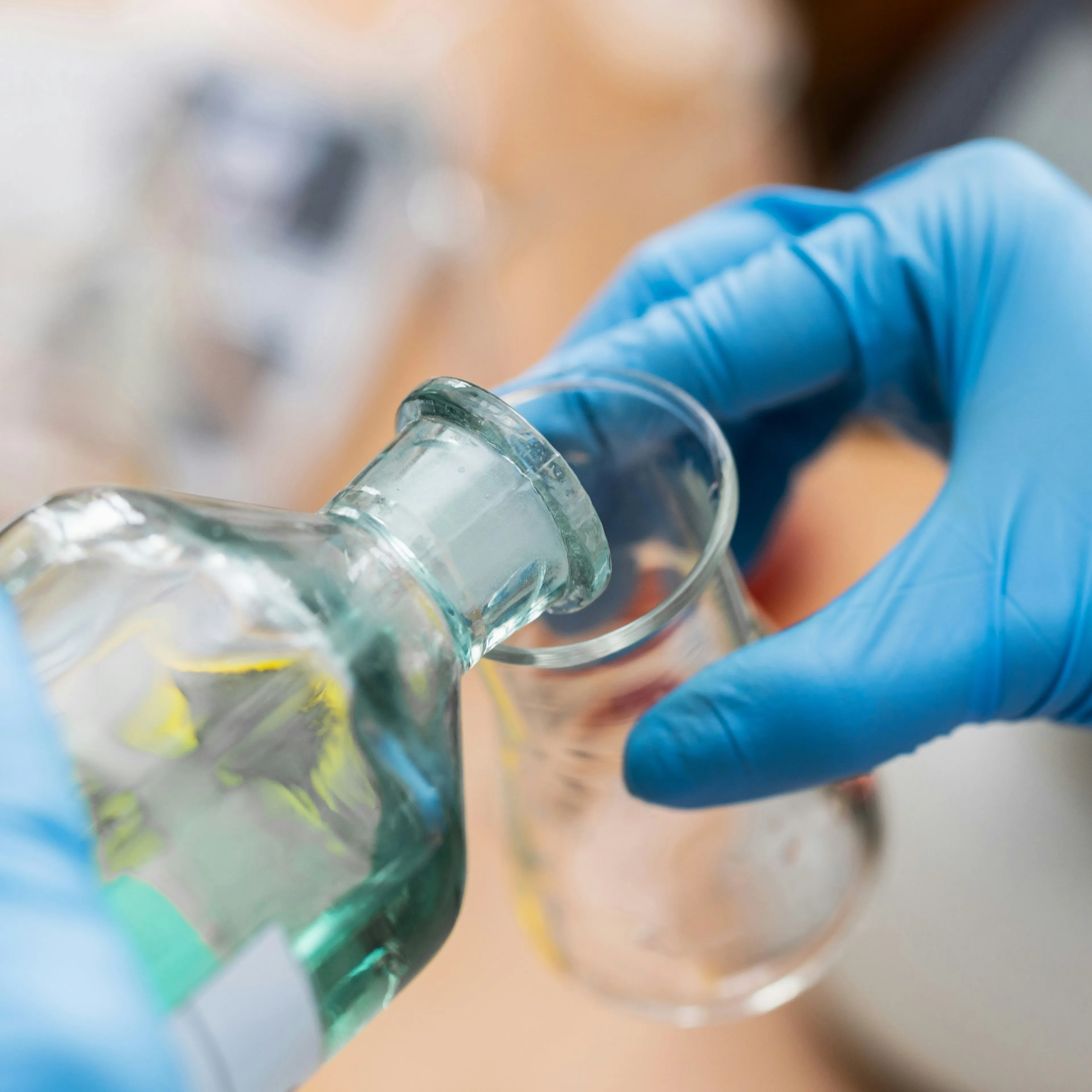 Gloved hands handling laboratory glassware with clear liquid in a scientific setting with blue gloves.