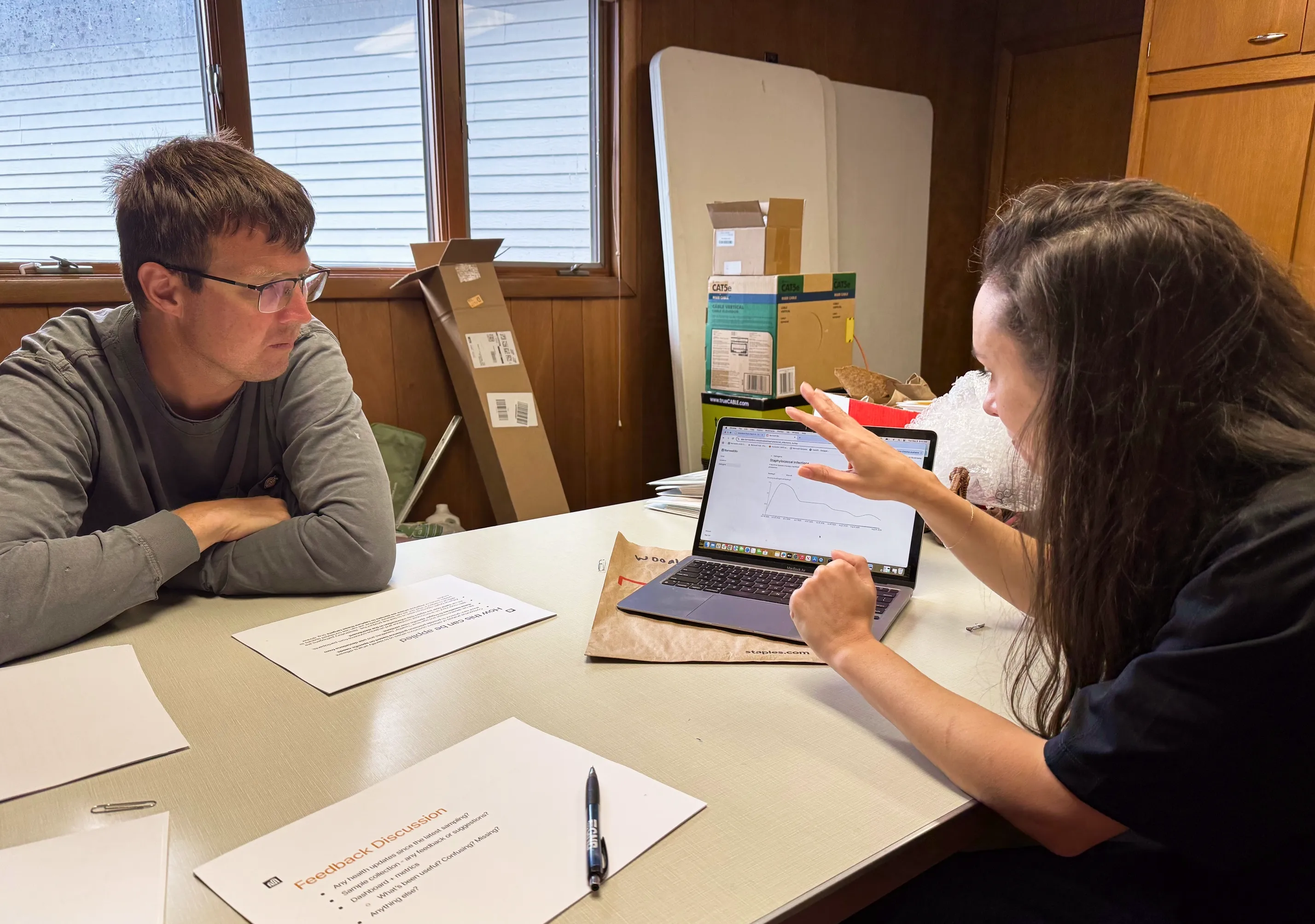 Two people at a table discussing data displayed on a laptop screen, surrounded by papers and cardboard boxes in a wood-paneled room.