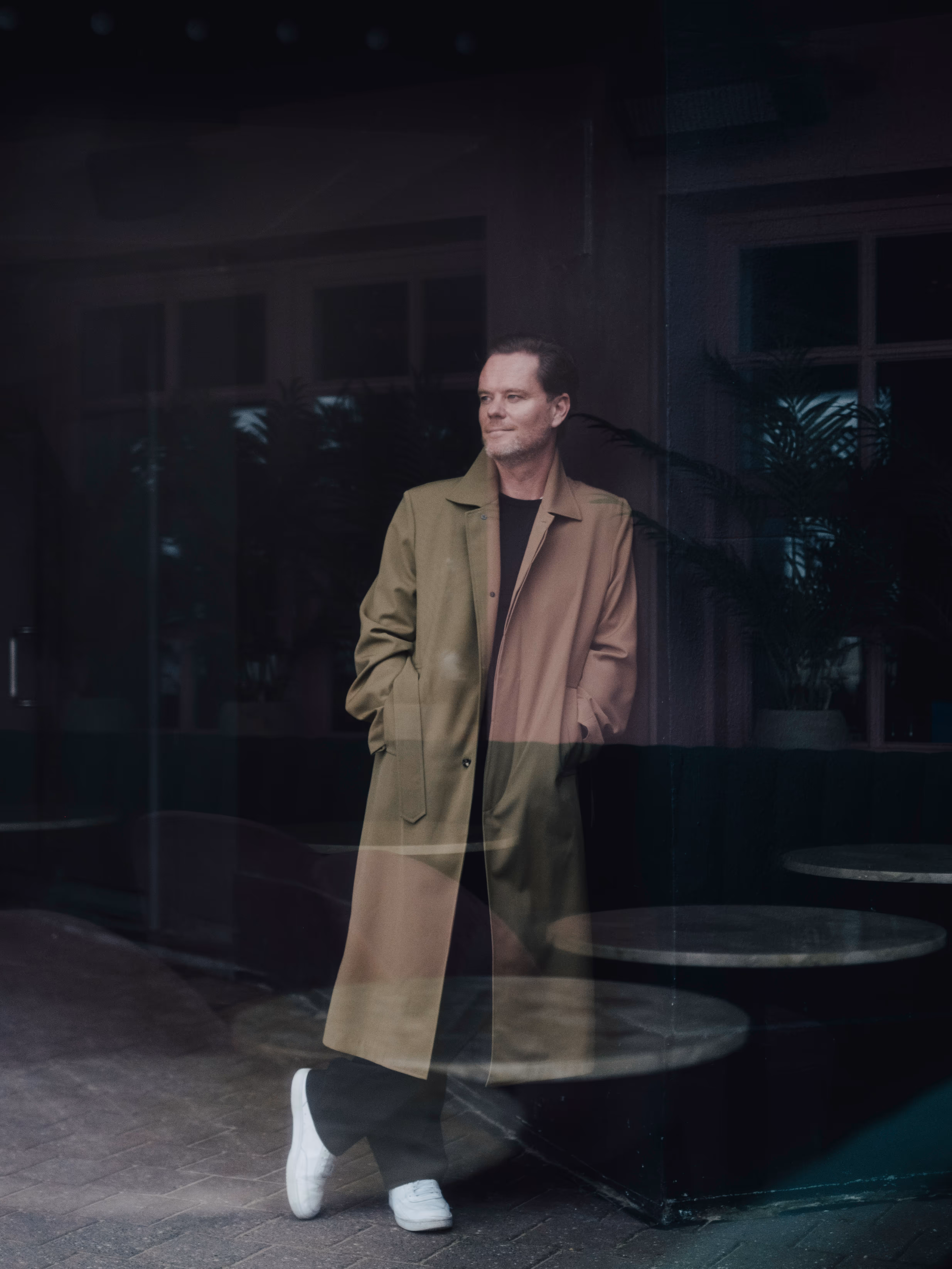 Tanguy in beige trench coat, black pants, and white sneakers standing with hands in pockets looking sideways, with indoor plants and tables behind glass.