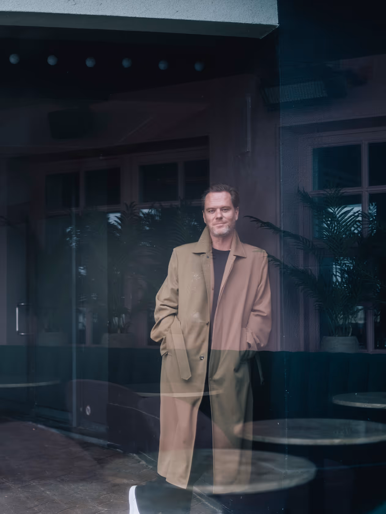 Tanguy wearing a long beige coat standing indoors behind glass with reflections of tables and plants.