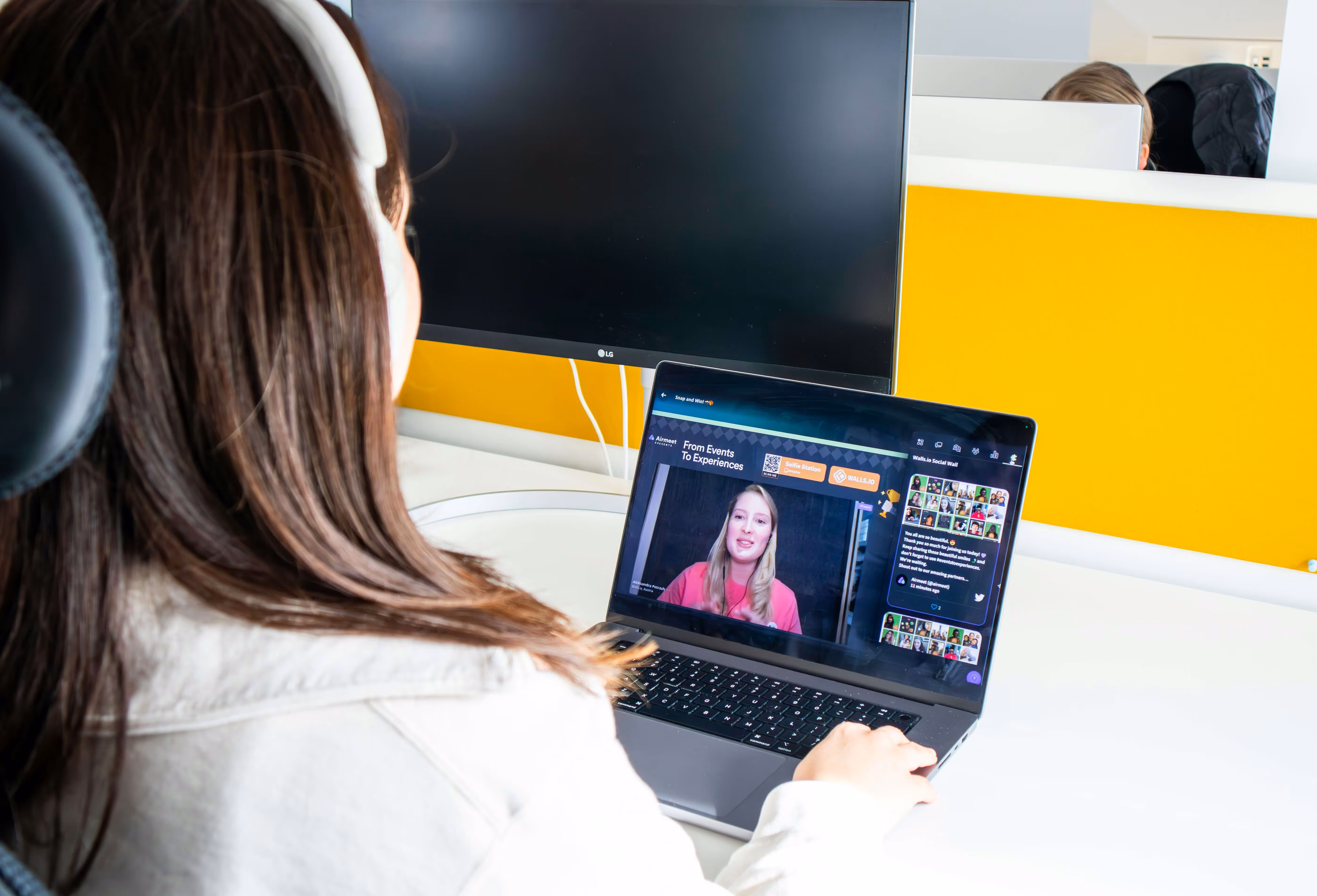 Person wearing white headphones attending a virtual meeting on a laptop in a bright office with yellow partitions.